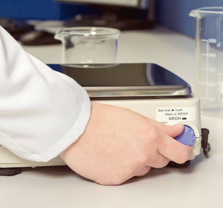 A hand turns the blue button of a laboratory device, possibly a balance, on a table with beakers in the background.