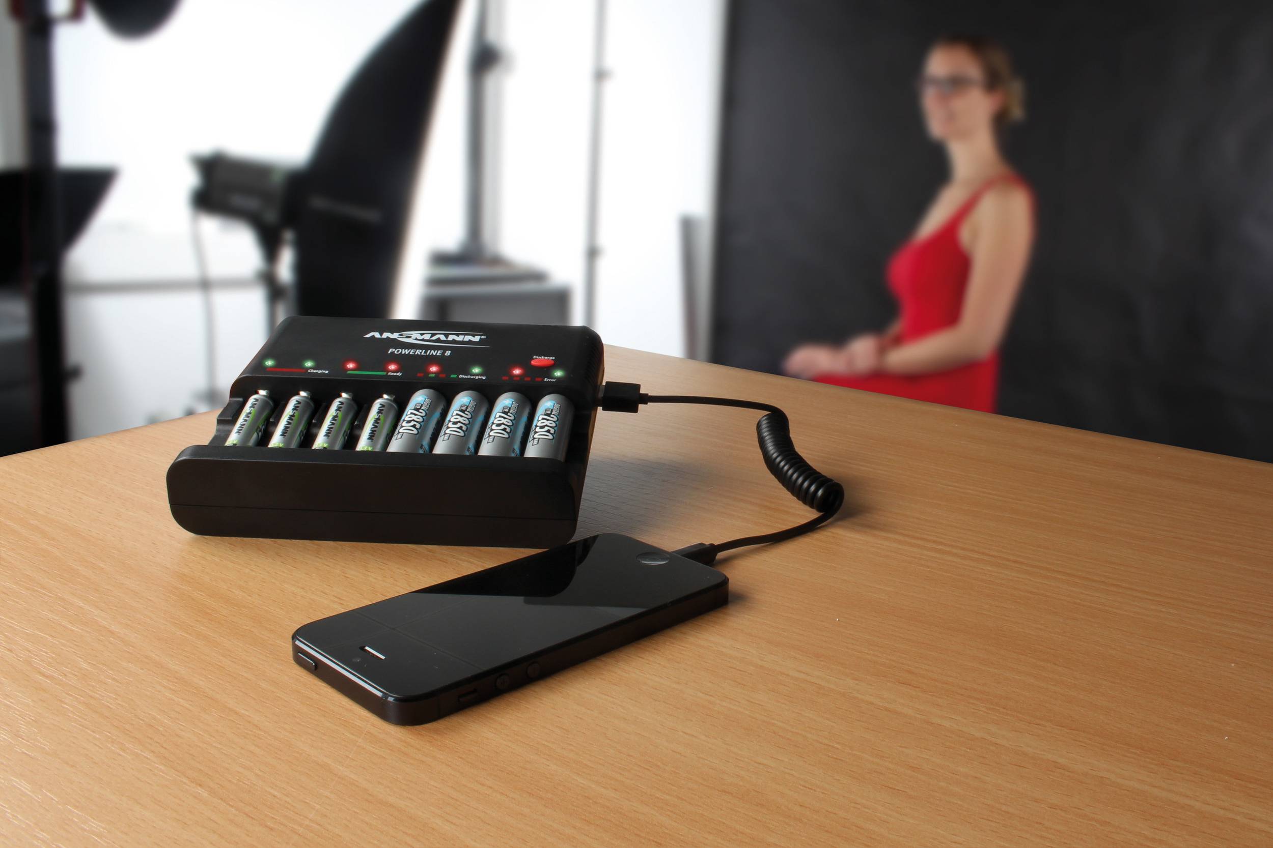 A battery charging station that is charging batteries is connected to a smartphone. A blurred person is standing in the background.