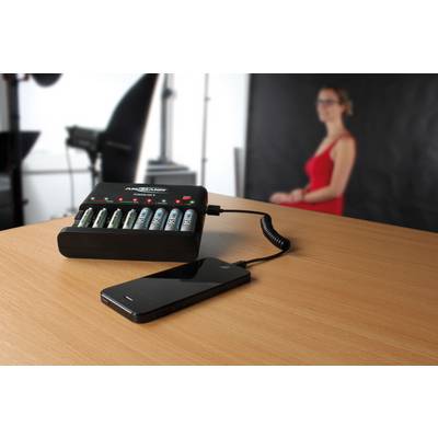 A battery charging station that is charging batteries is connected to a smartphone. A blurred person is standing in the background.