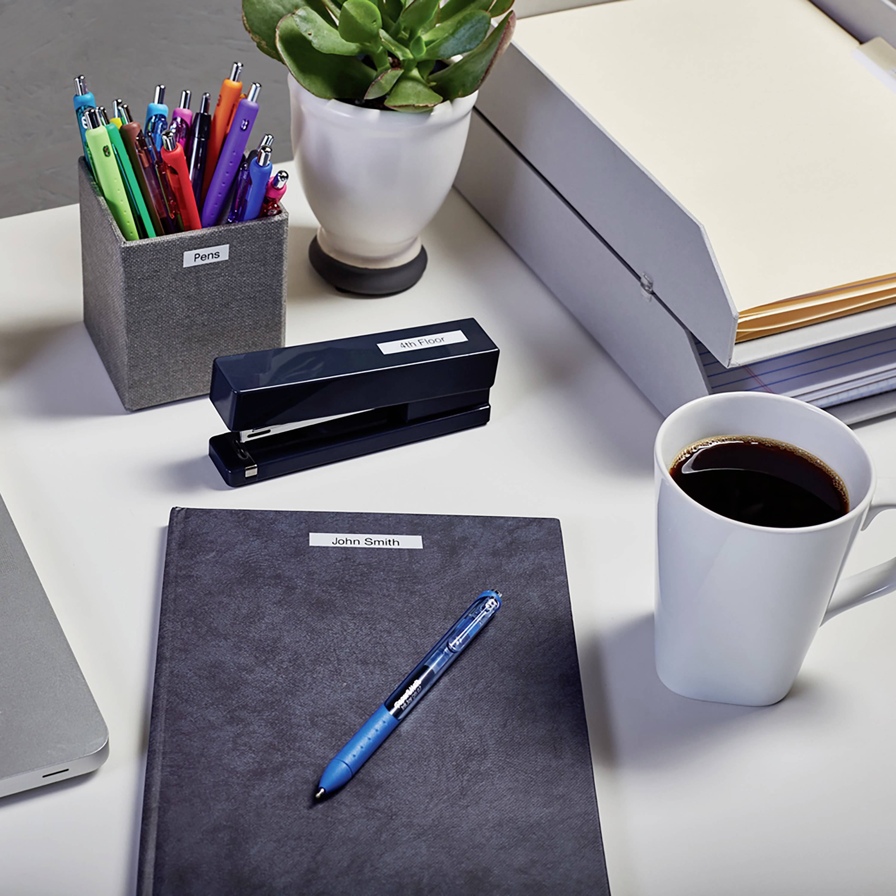 A desk with pens in a pen holder, a notebook, a cup of coffee, a stapler and plants in the background.