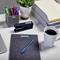 A desk with pens in a pen holder, a notebook, a cup of coffee, a stapler and plants in the background.