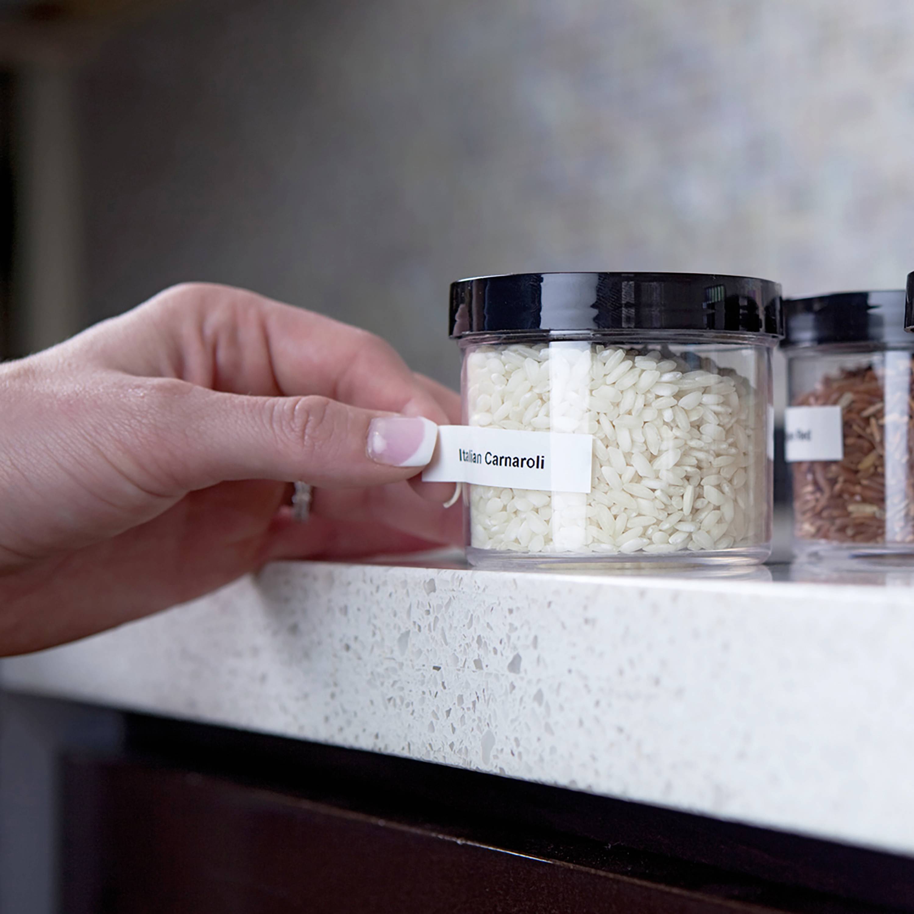 A hand reaches for a glass container labelled 'Riso Carnaroli'. In the background, additional containers of rice are visible.