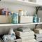 Shelves with cleaning and household items, including glass containers with labels, cleaning products, towels, and detergent boxes.