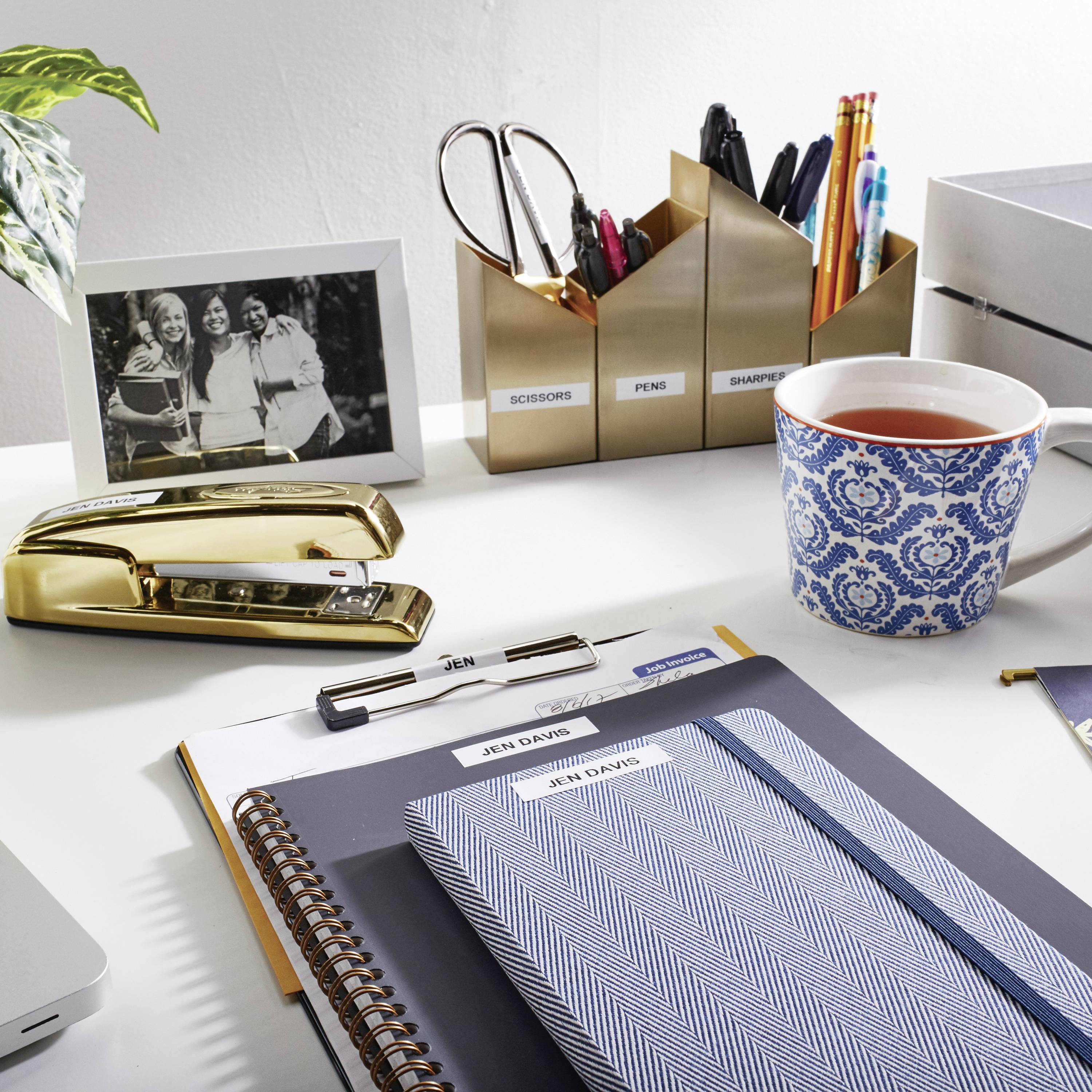A tidy desk with a cup of tea, pencil holders, a notebook and a black-and-white framed photograph of three people laughing.
