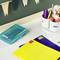 Office supplies on a desk: blue and yellow folders, coloured pens in a container, a box of labels, and bunting on the wall.