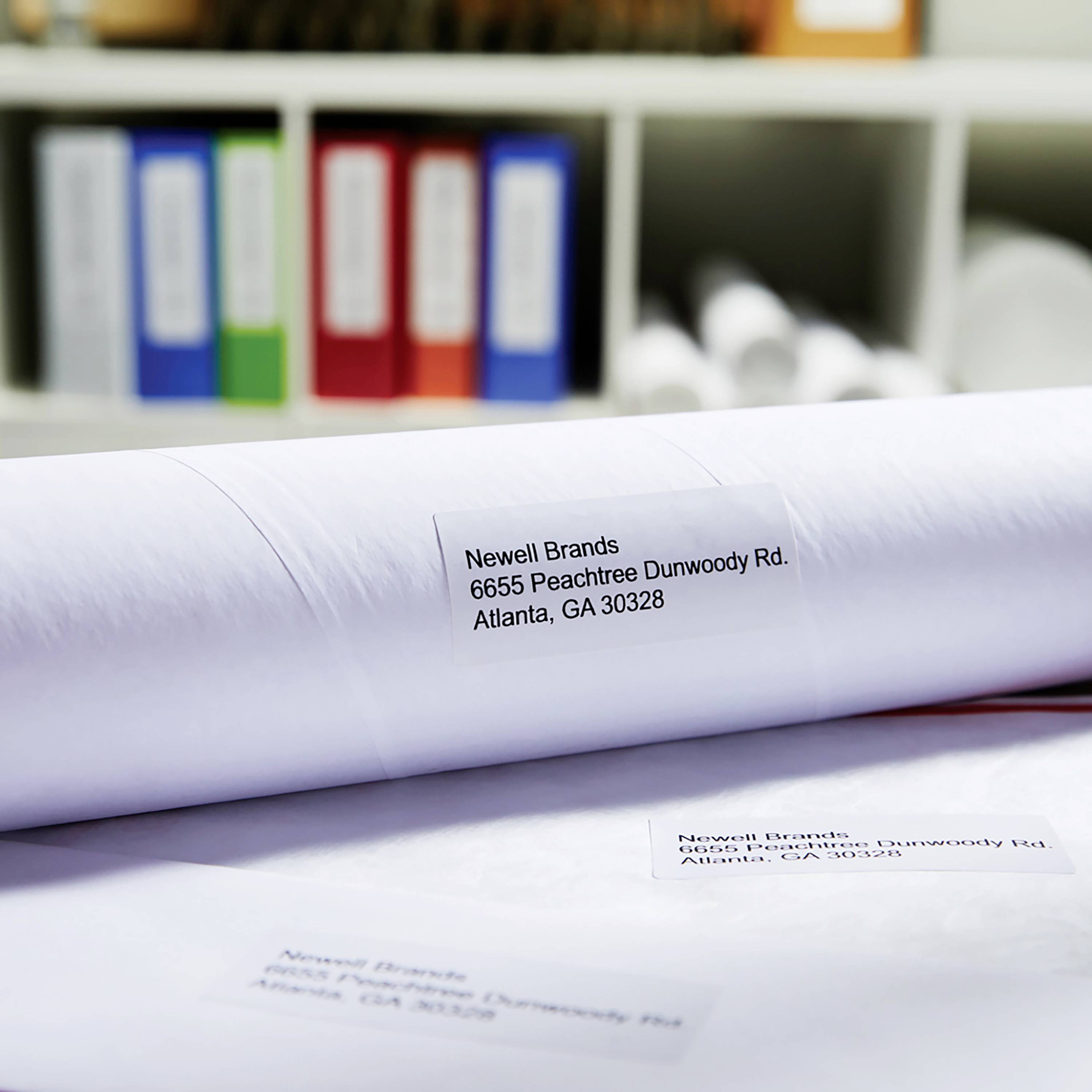 A roll with a label reading 'Newell Brands, 6655 Peachtree Dunwoody Rd, Atlanta, GA 30328' in front of blurred file folders in the background.