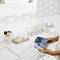 A person is operating a labelling device on a marble washbasin in the bathroom, surrounded by toothbrushes, soap dispensers, and glass containers.