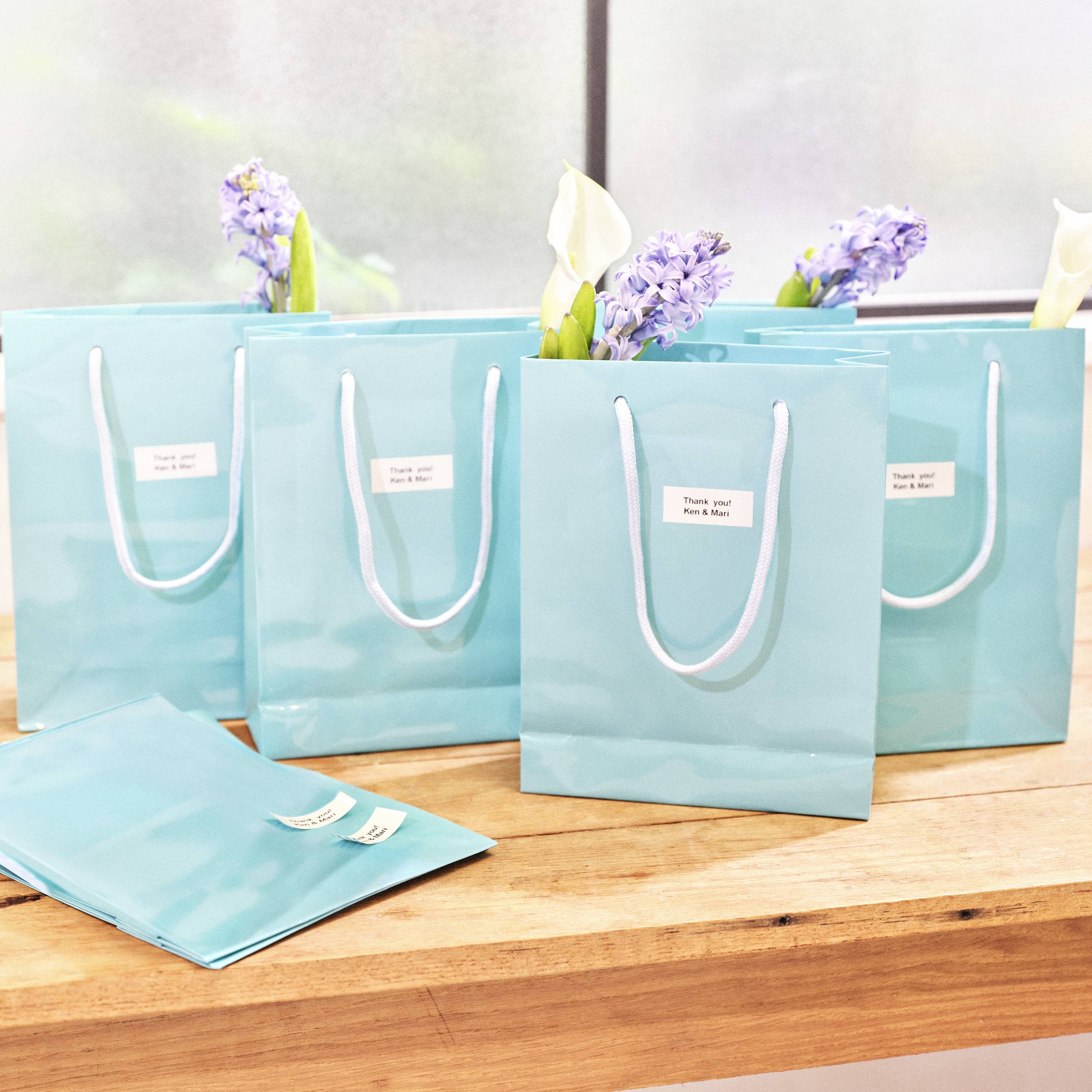 Turquoise gift bags decorated with flowers, placed on a wooden table in a bright setting.