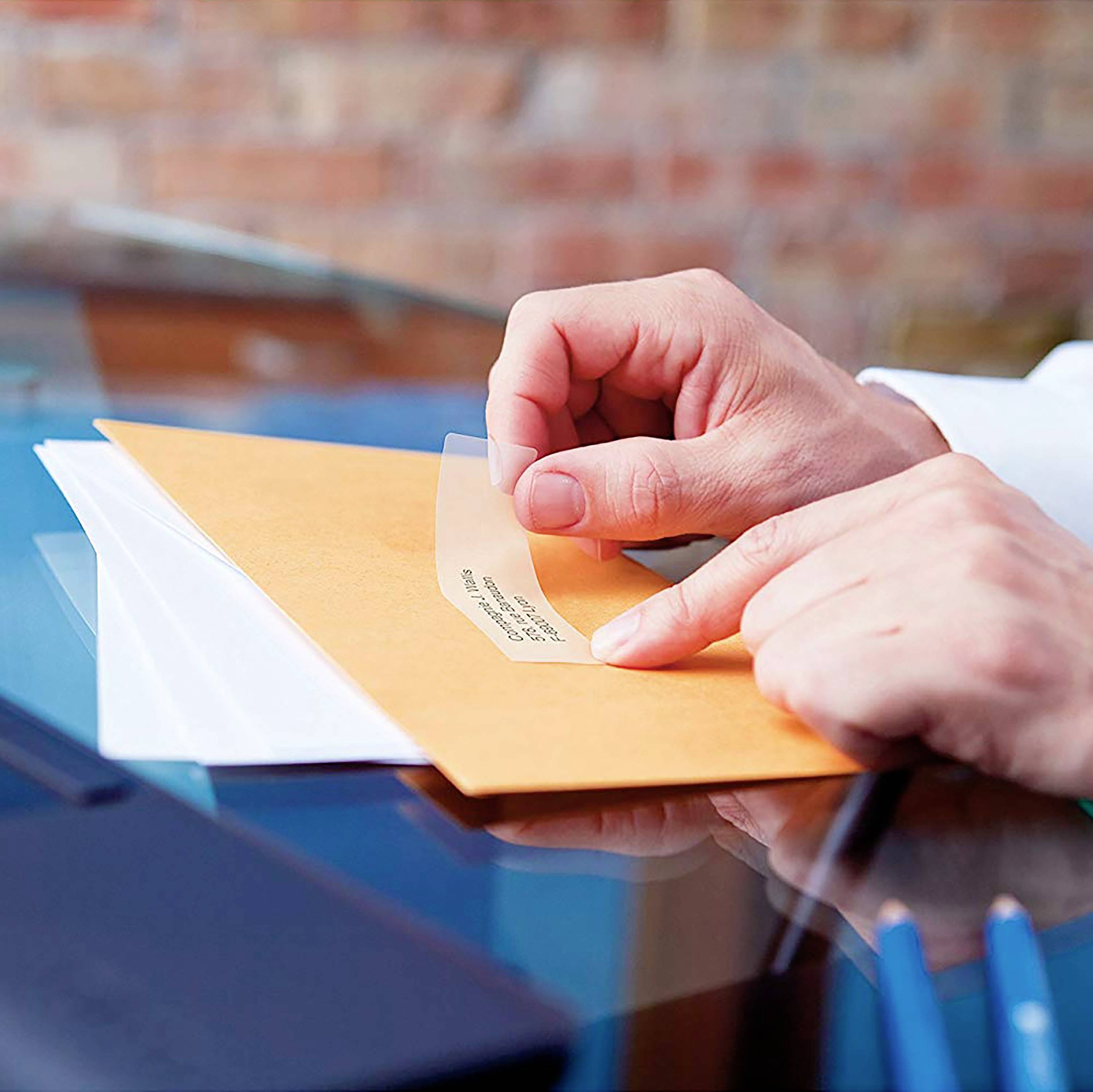 A person is sticking a transparent label onto a yellow envelope. Other documents are lying on the table.