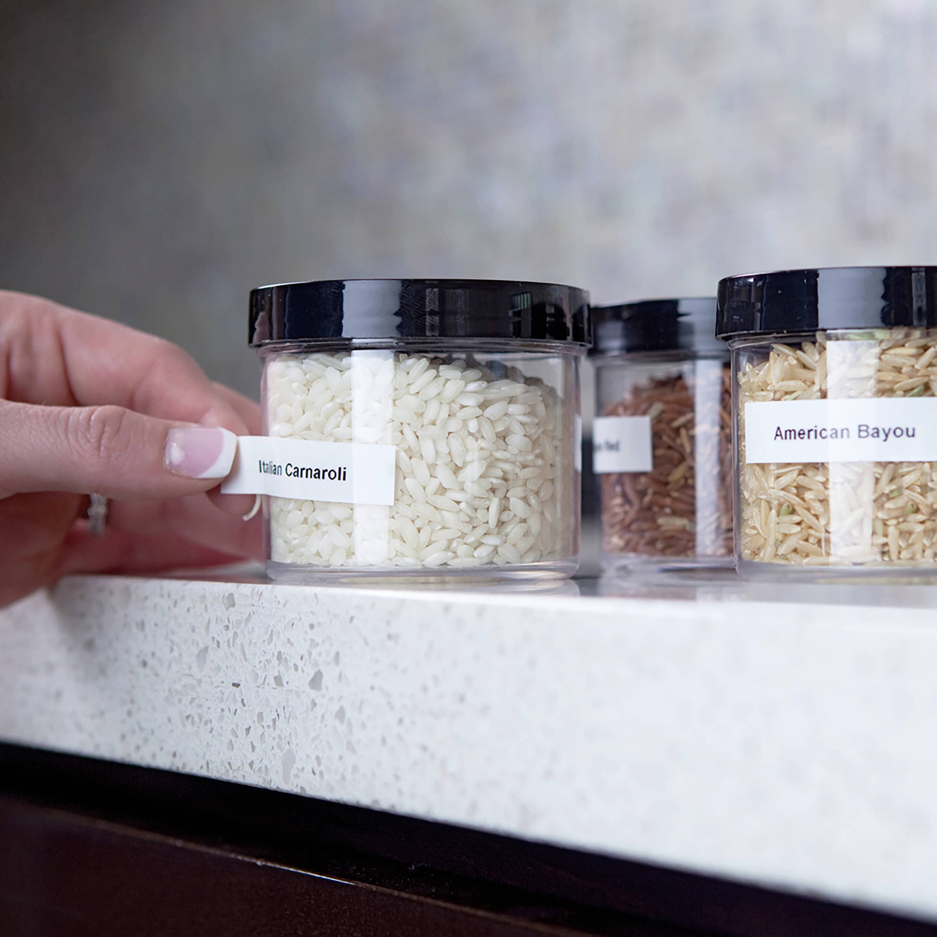 A hand reaches for a glass of Italian Carnaroli rice on a worktop. Beside it are glasses containing various types of rice.
