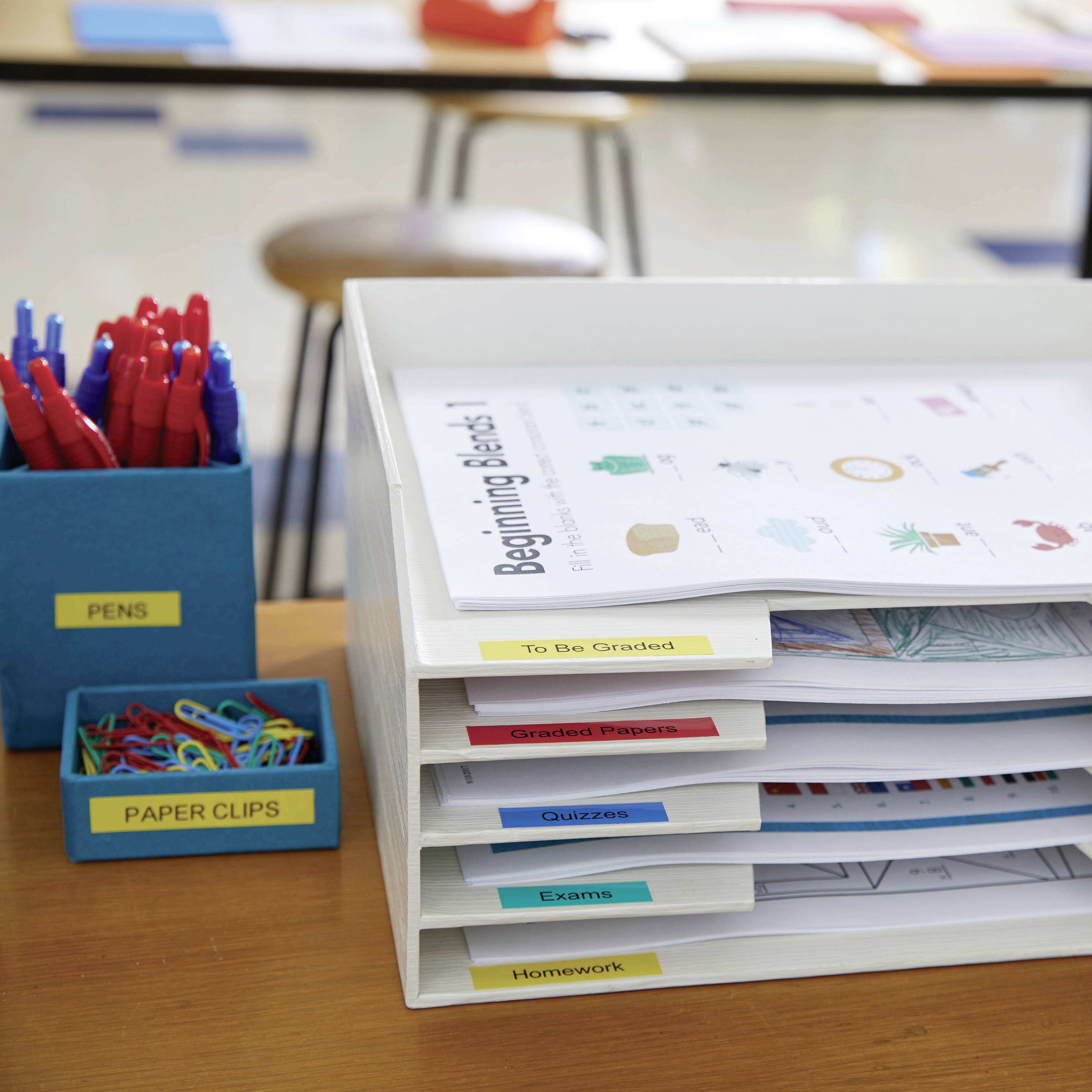 A desk organiser with labelled compartments: 'To Be Graded', 'Graded Quizzes', 'Exams', 'Homework'. Next to it are pens and paperclips.
