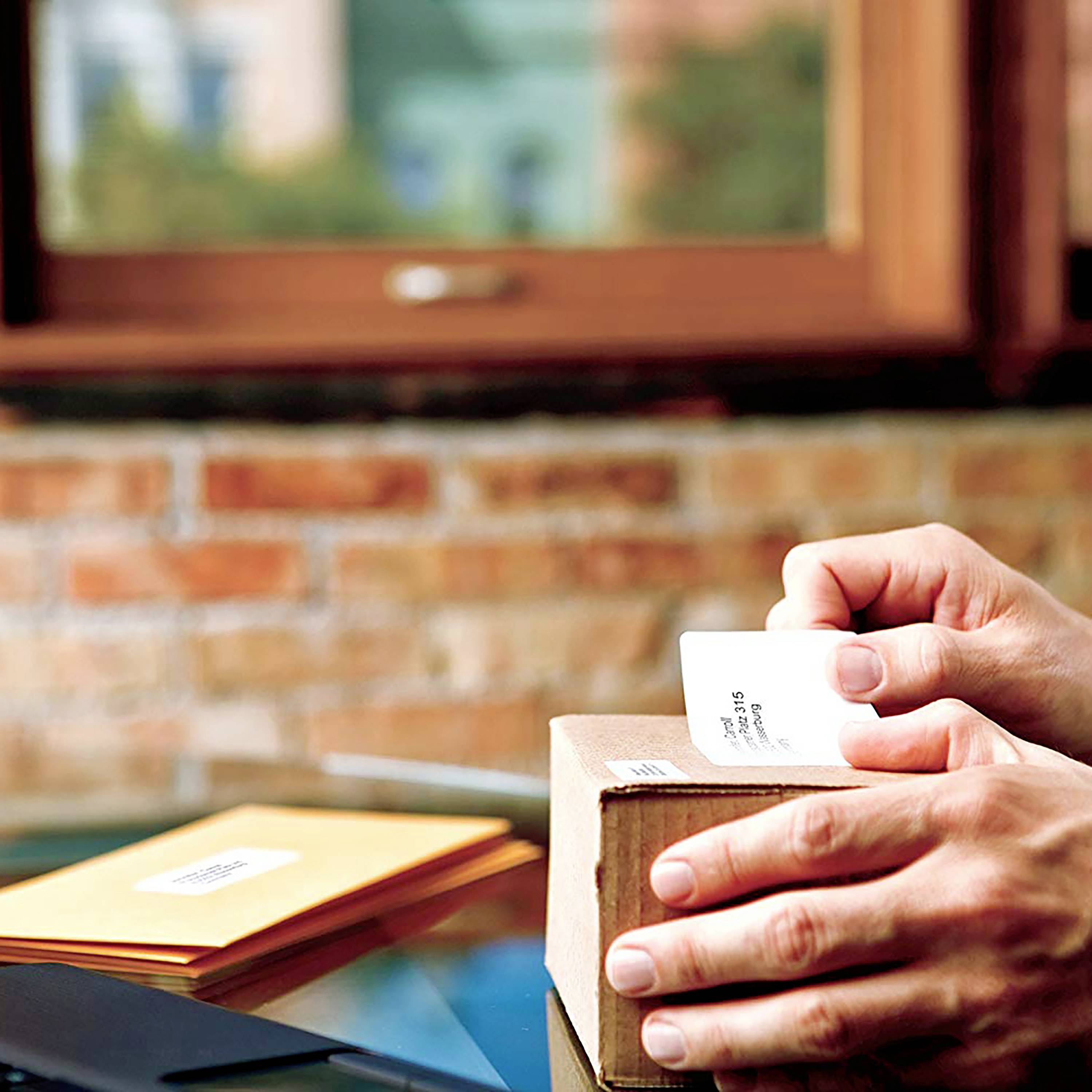 A person is holding a small, brown parcel, ready for posting or delivery. Envelopes are visible in the background.