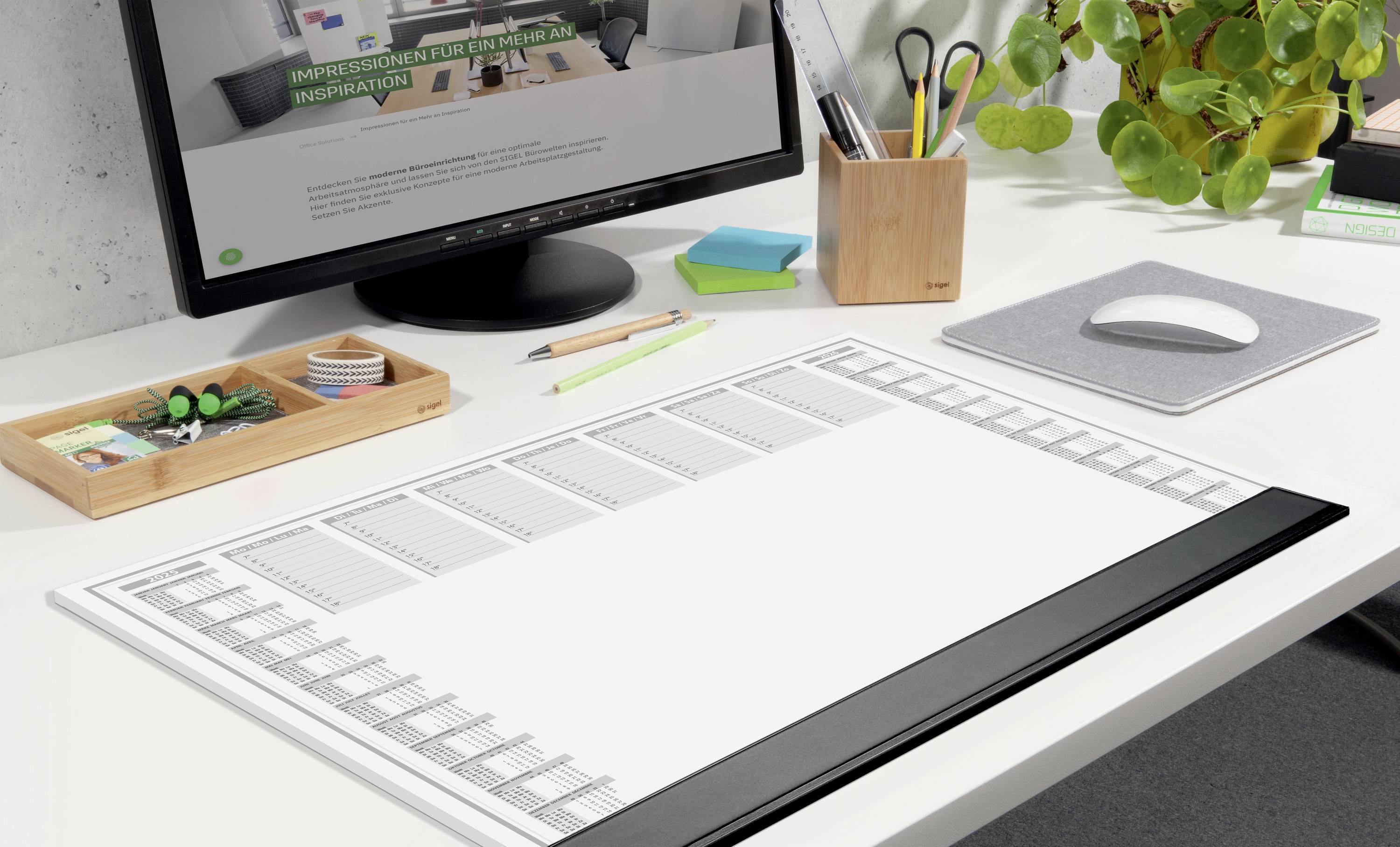 Desk with a large desk calendar, surrounded by folders, pens and a plant. In the background, a computer screen.