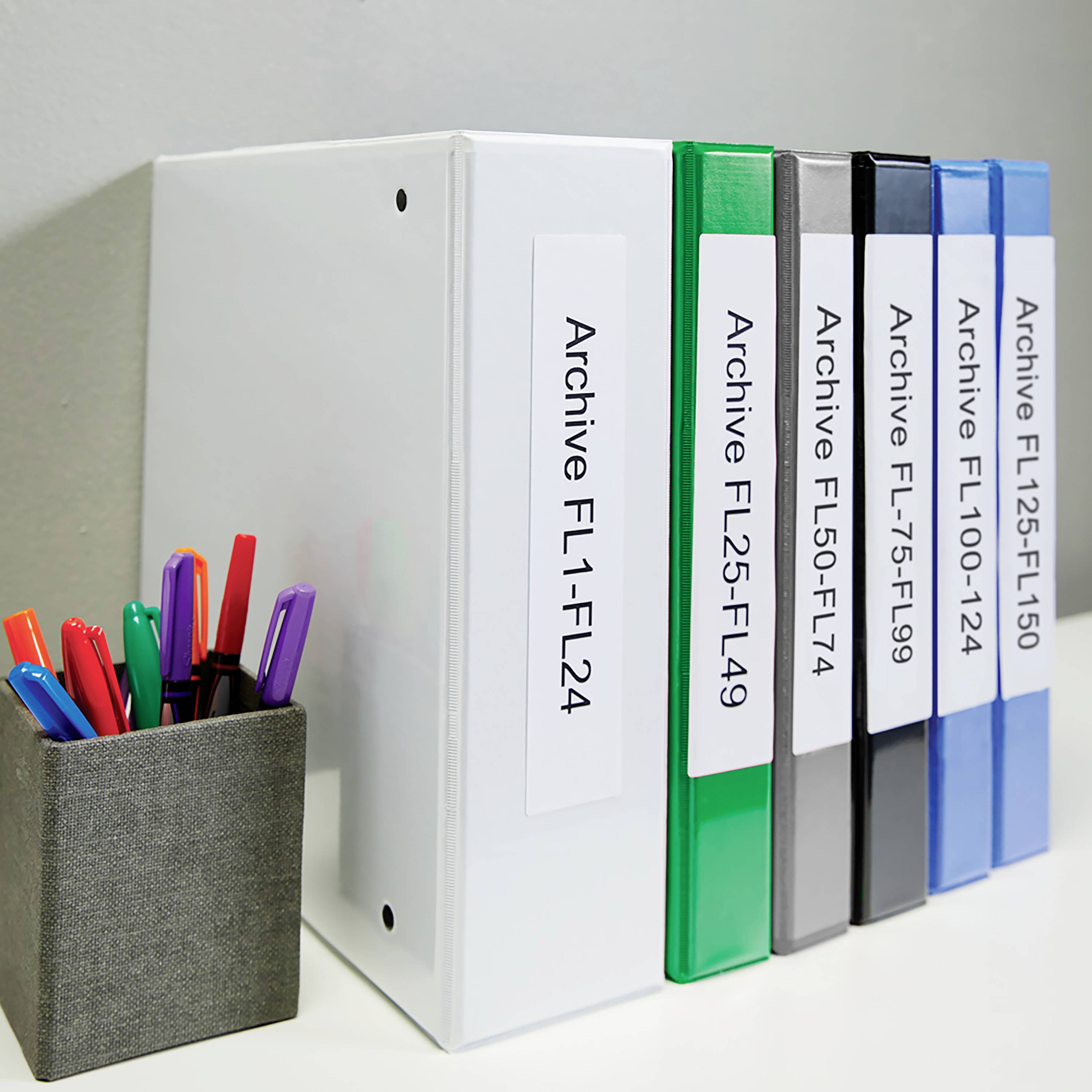 Folders in various colours with labels 'Archive FL1-L25' through to 'FL125-L150' are lined up side by side. Beside them is a container of pens.