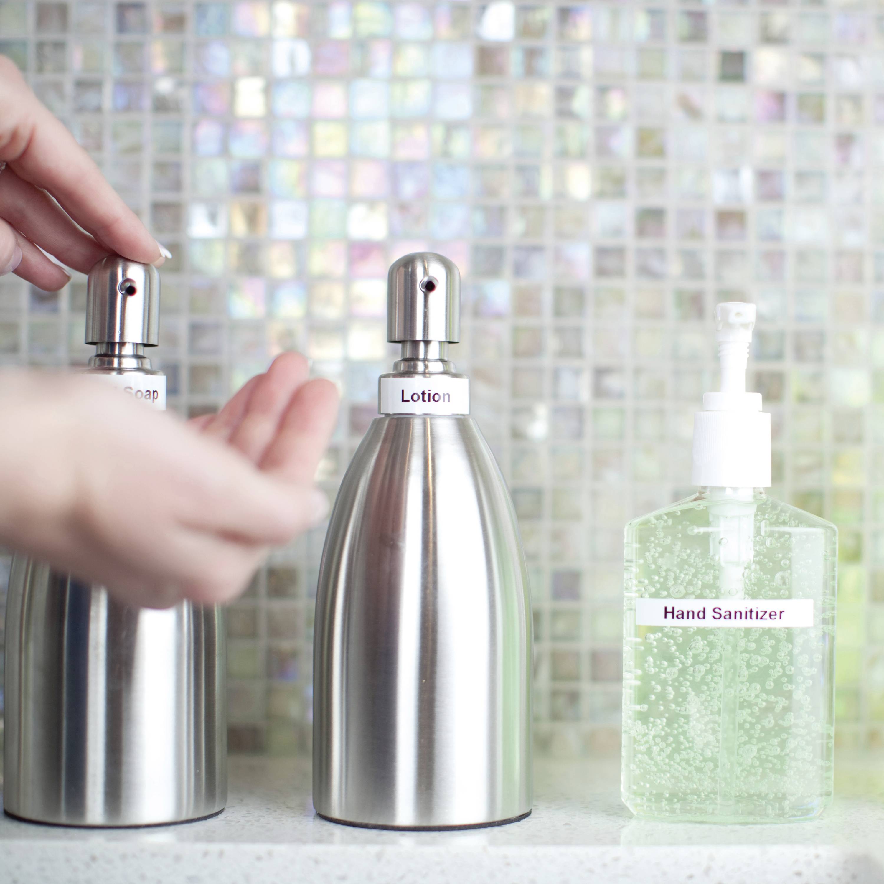 A hand presses a soap dispenser next to a lotion dispenser and a hand sanitiser on a patterned surface.