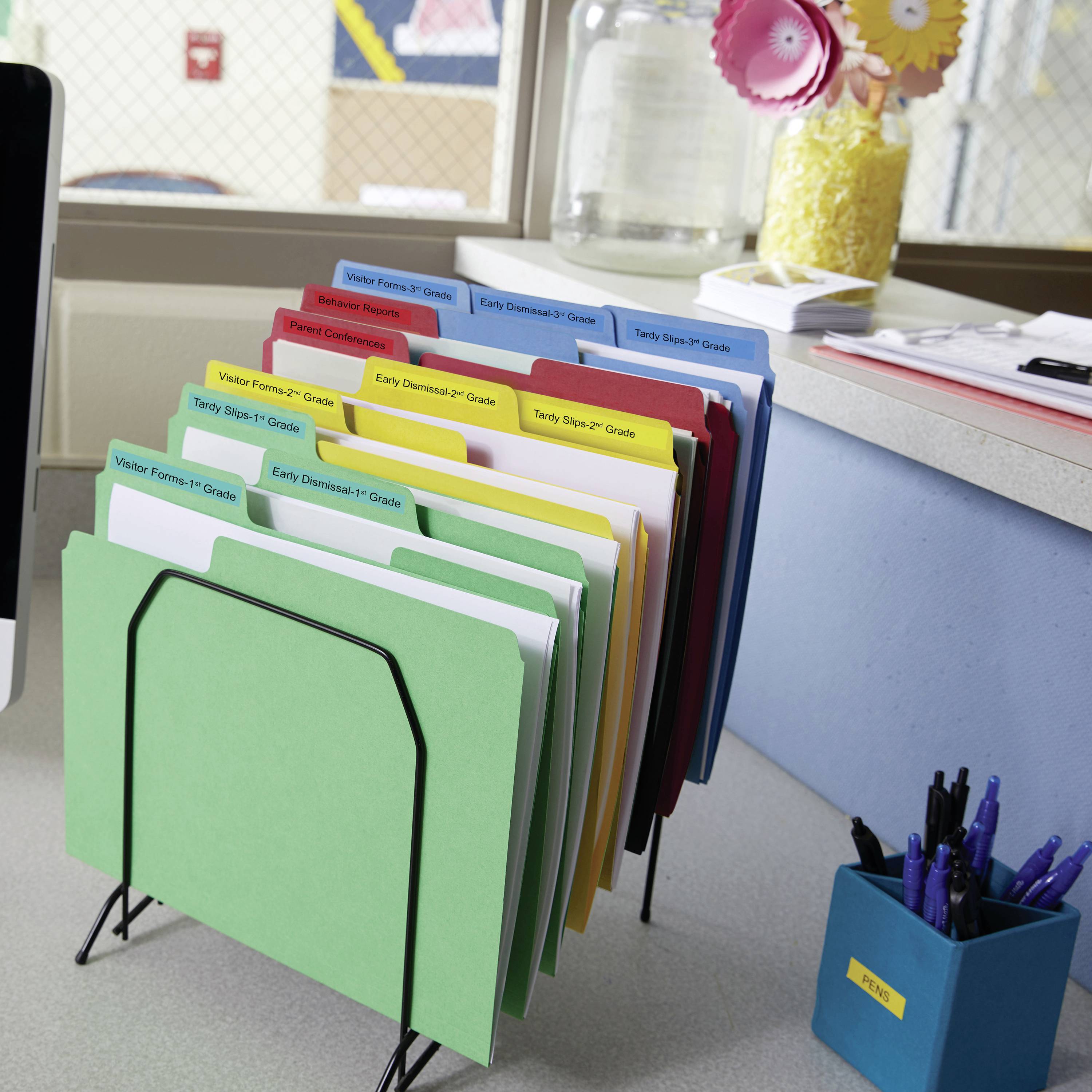 A desk with a file holder containing colourful folders labelled for different school year groups. In the background, a glass vase with decorative flowers.