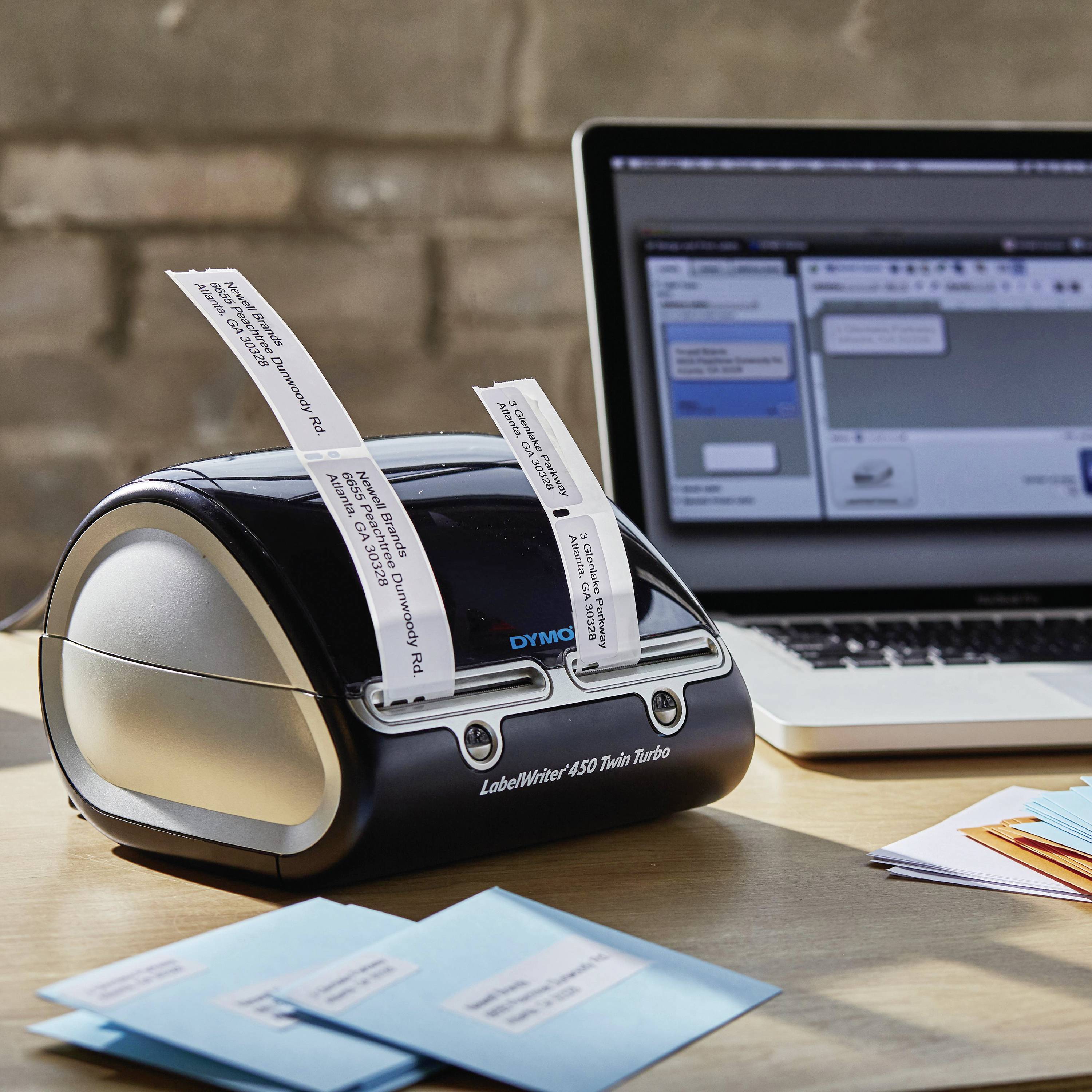 A label printer sits on a table beside a laptop. The printer is producing printed labels while the laptop displays a software application.