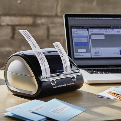 A label printer sits on a table beside a laptop. The printer is producing printed labels while the laptop displays a software application.