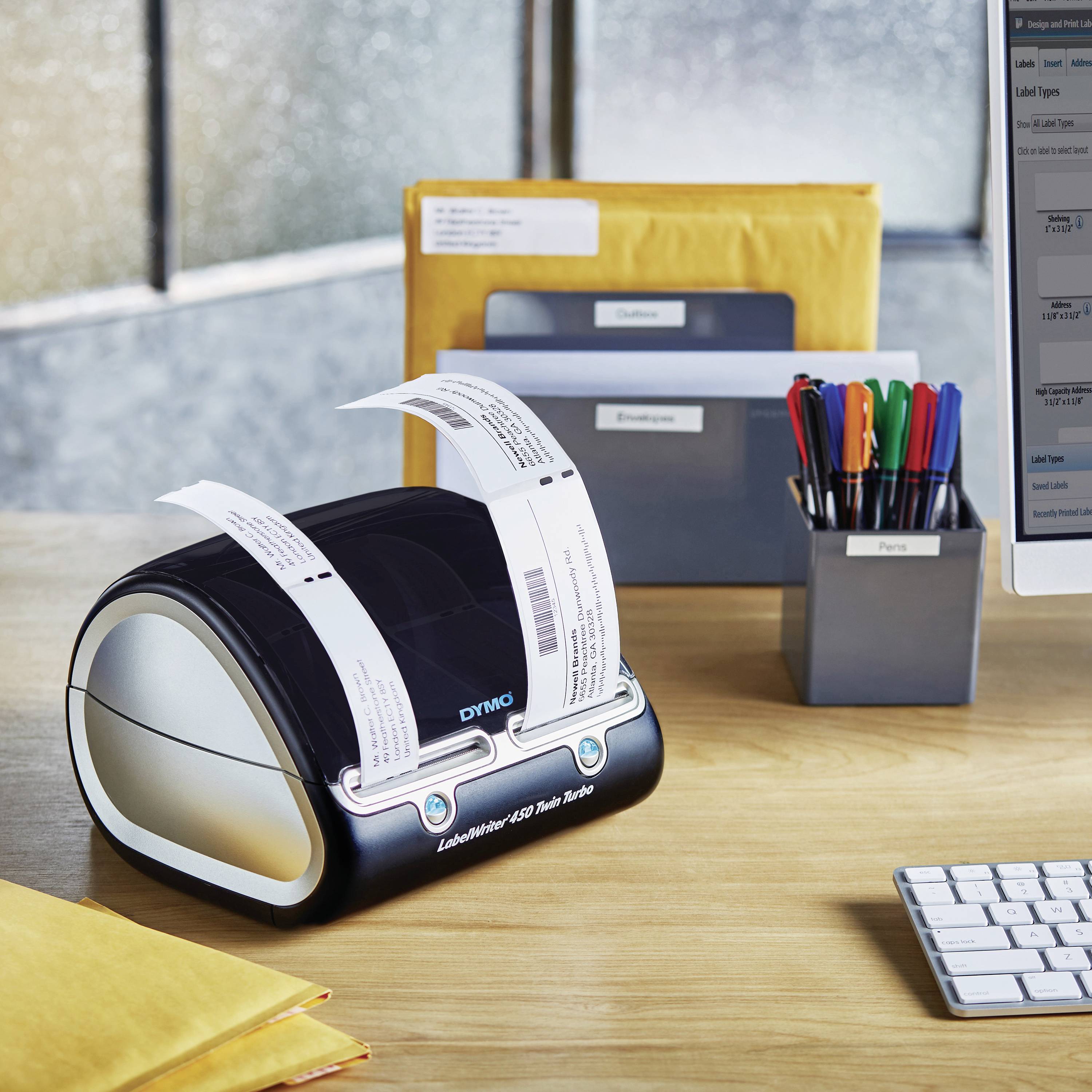 A label printer sits on a desk, printing a label. In the background, writing materials, envelopes, and a monitor are visible.