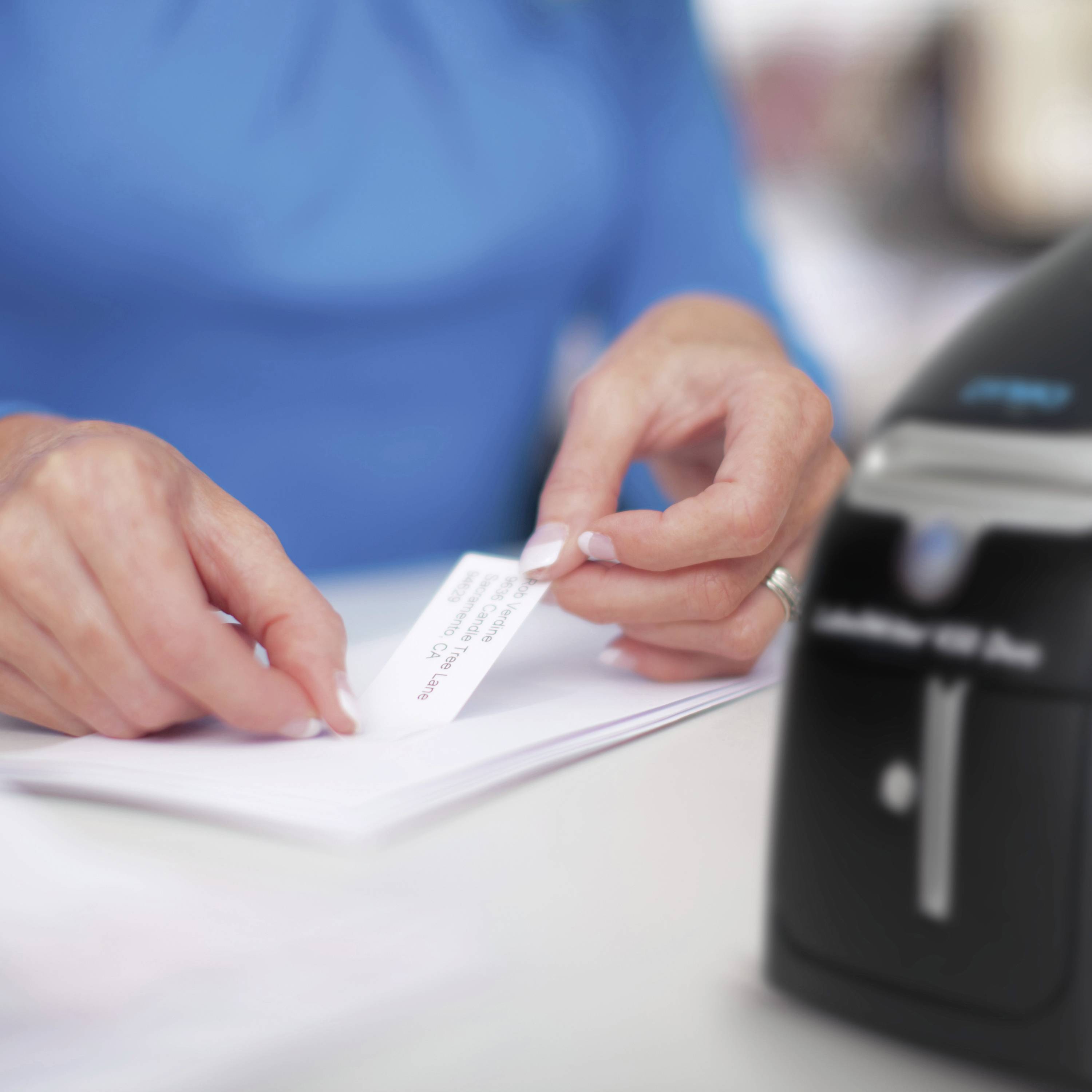 A person inserts a label with the headings 'Name' and 'Address' into a labelling machine. The machine is standing on a table.
