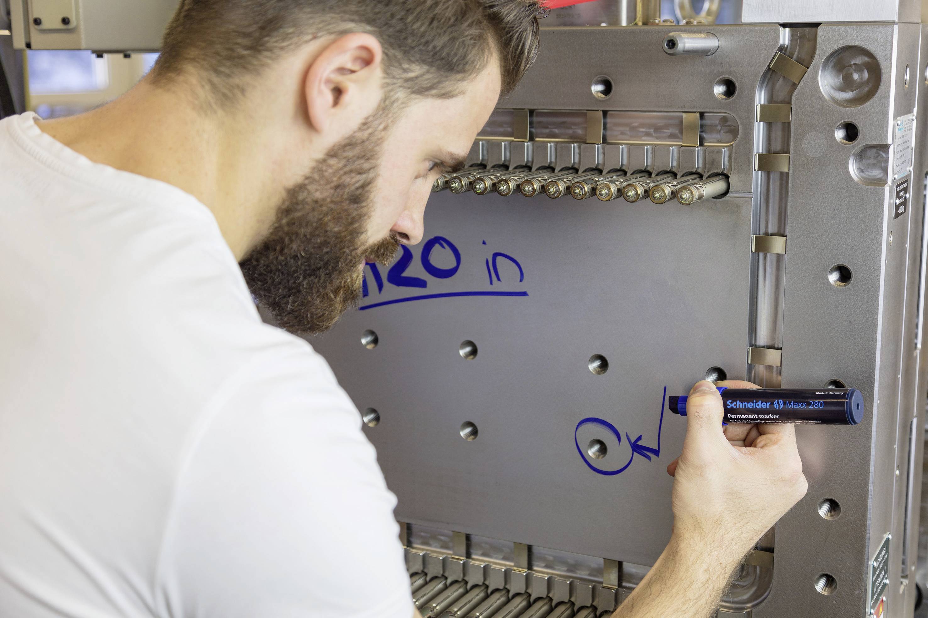 A man in a white T-shirt is writing numbers and letters with a blue marker on a metallic surface with technical elements.
