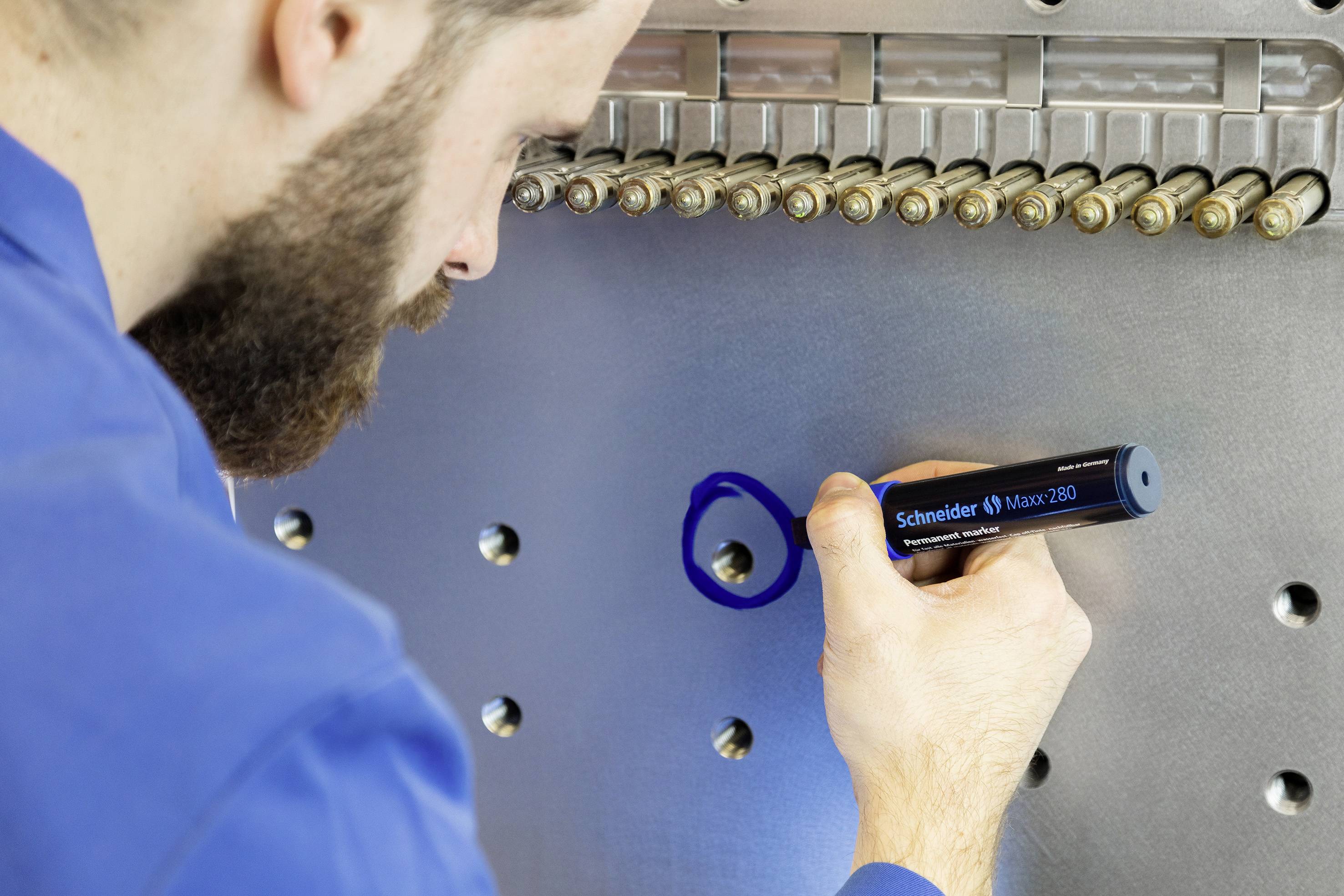 A man in blue workwear is marking a metal surface with a blue pen. Several rivets or bolts are visible.