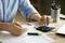 A person is calculating on a calculator and checking documents at a desk. A mug filled with pens sits nearby.