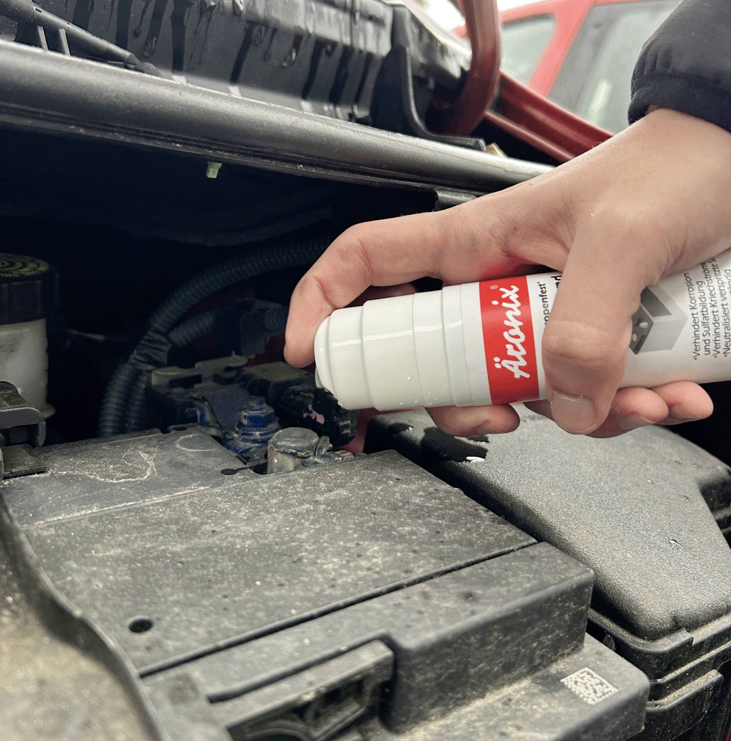 A hand sprays a cleaning solution onto the car battery terminal under the bonnet of a car.