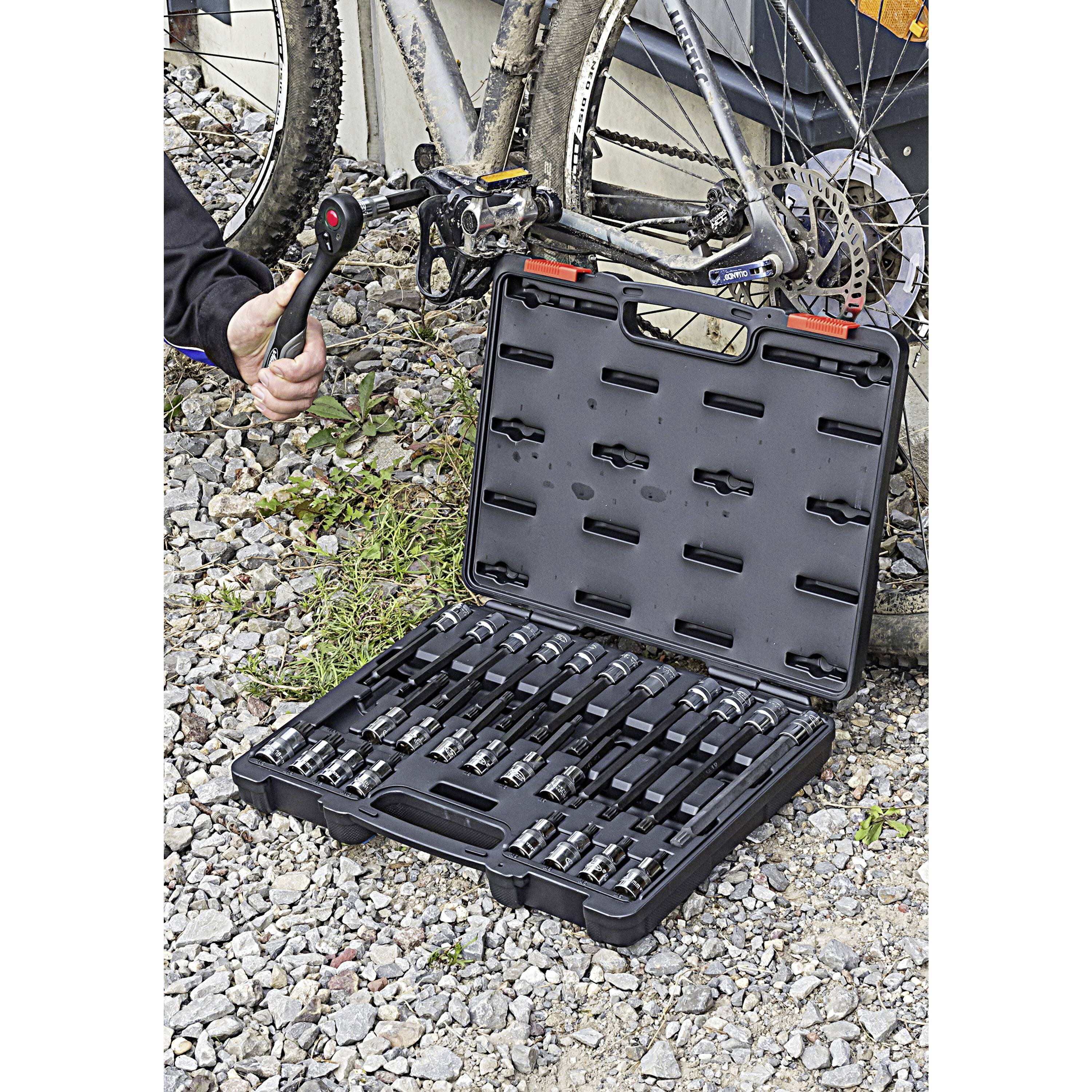 A person uses a tool from a bicycle tool kit, which is open on the gravel next to a mountain bike.