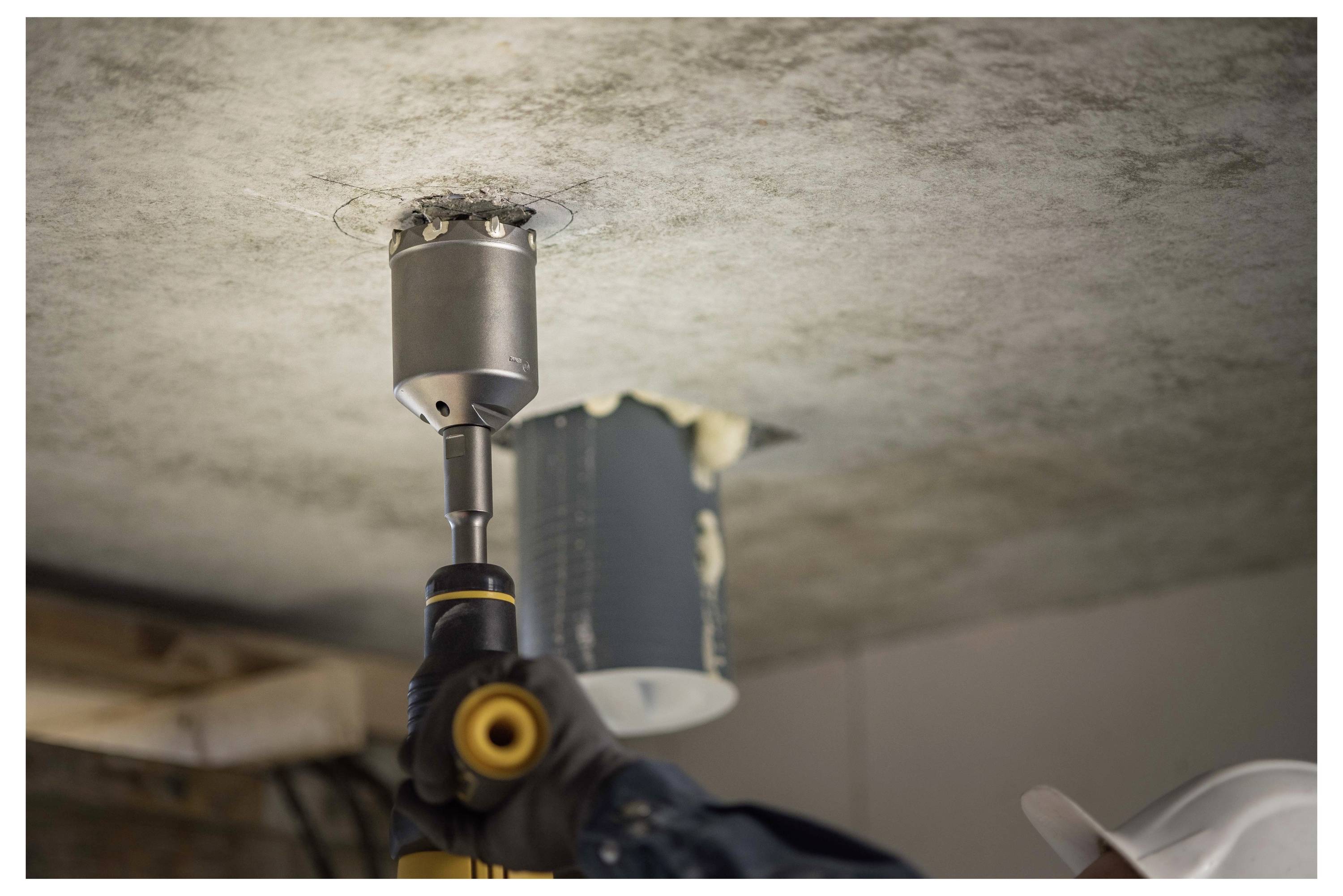A worker uses a hole saw drill to cut a circular hole in a ceiling, likely for installing a recessed light or fixture.