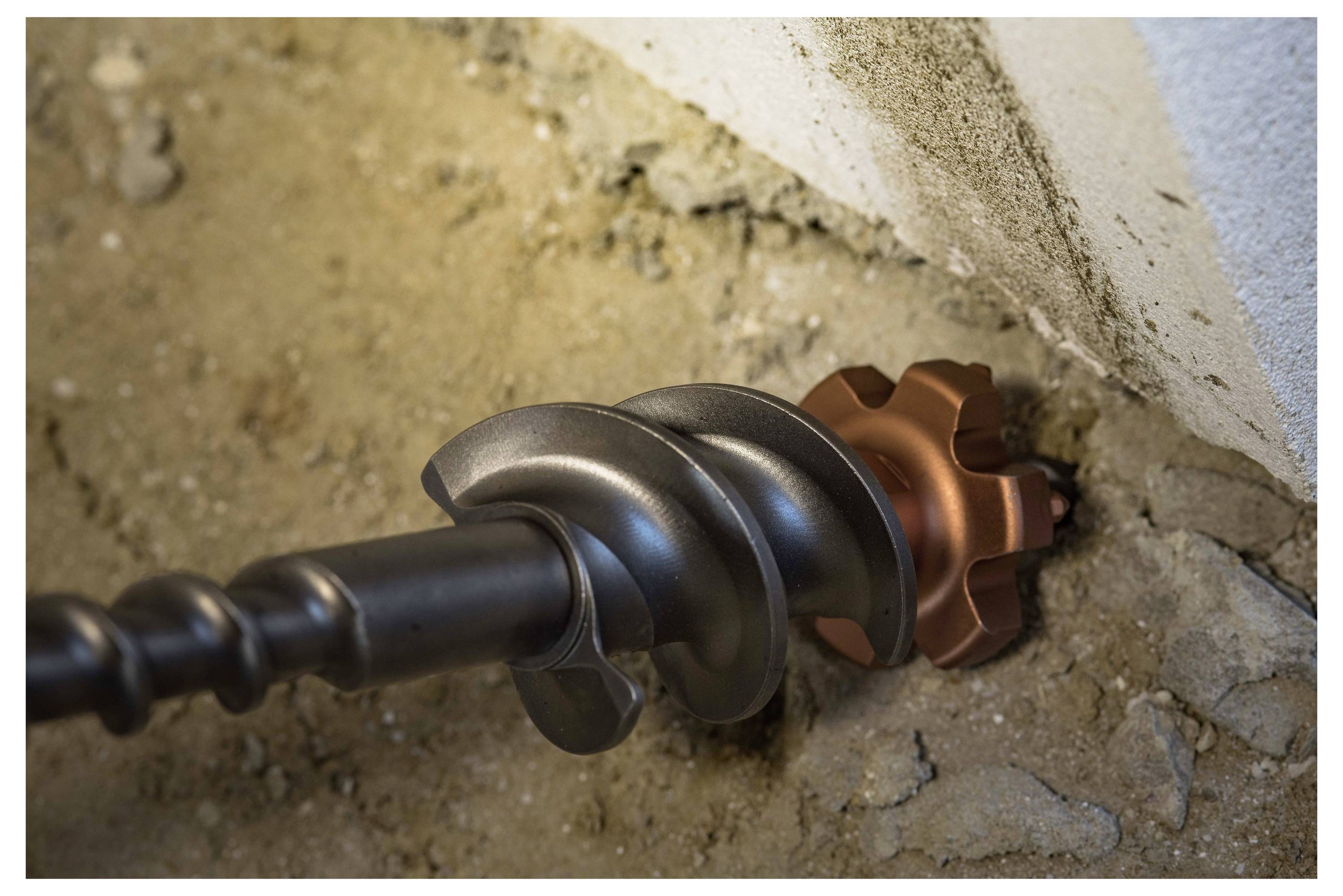 Close-up of a drill bit boring into a wall, with dust and exposed concrete visible around the hole.