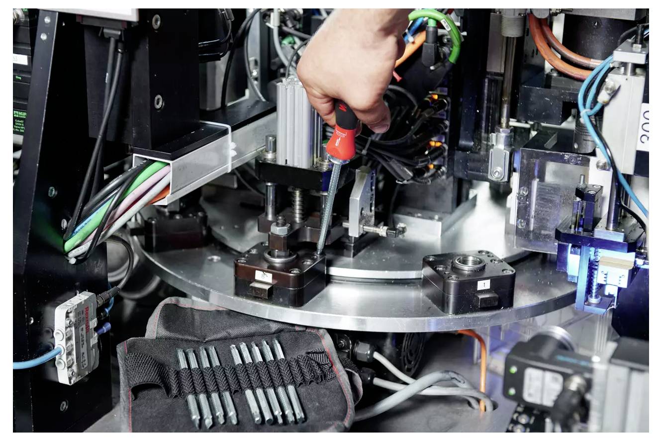 'A person uses a screwdriver to adjust machinery, surrounded by various mechanical components and cables in an industrial setting.'