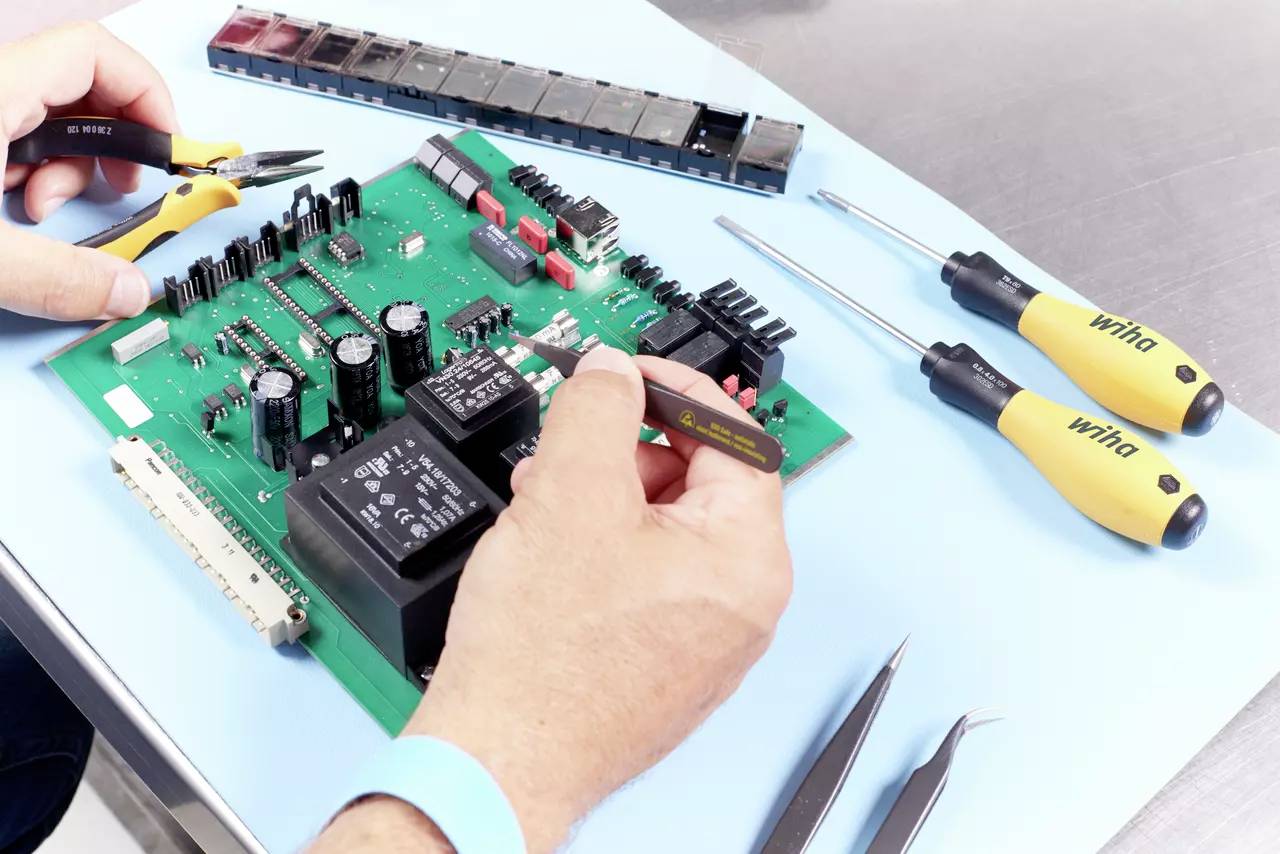 A person is repairing a green circuit board with tools on a table.