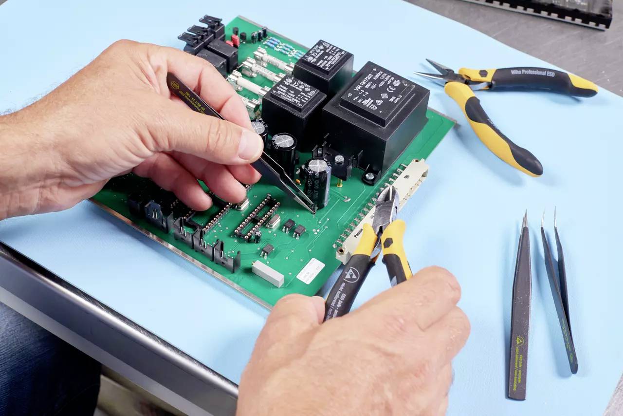 A person is repairing a circuit board with tweezers and side cutters. Tools are scattered across the table.