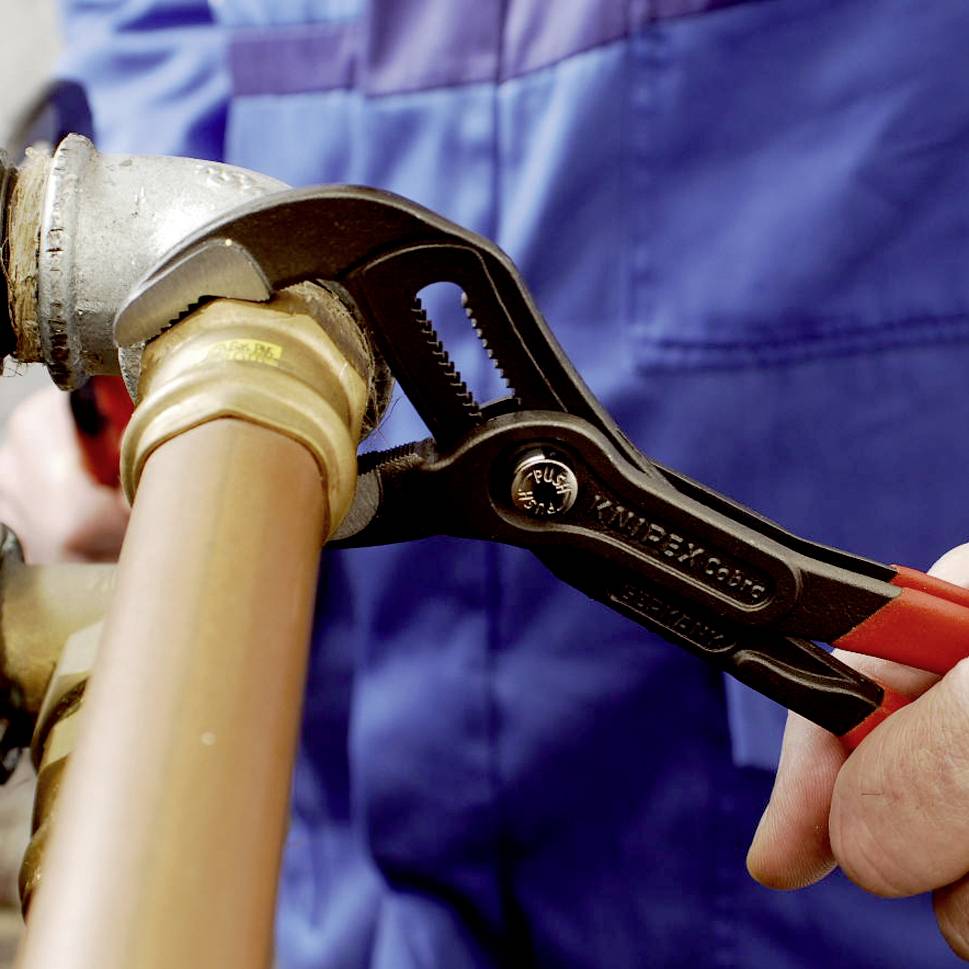 A person wearing blue workwear is repairing a copper pipe with a red adjustable spanner.