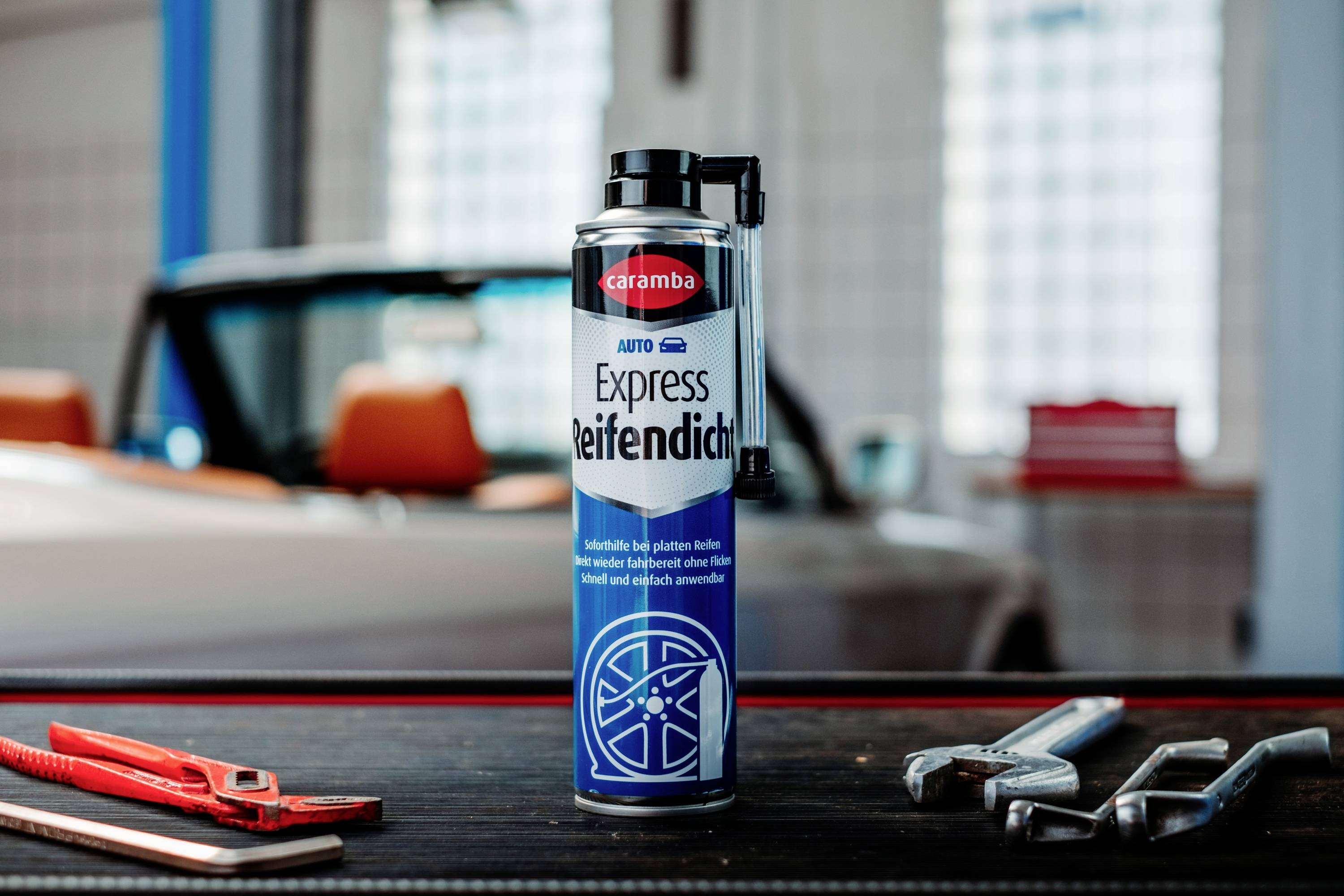 A can of 'Caramba Express Reifendicht', a tire sealant, on a table with wrenches and pliers, blurred classic car in the background.