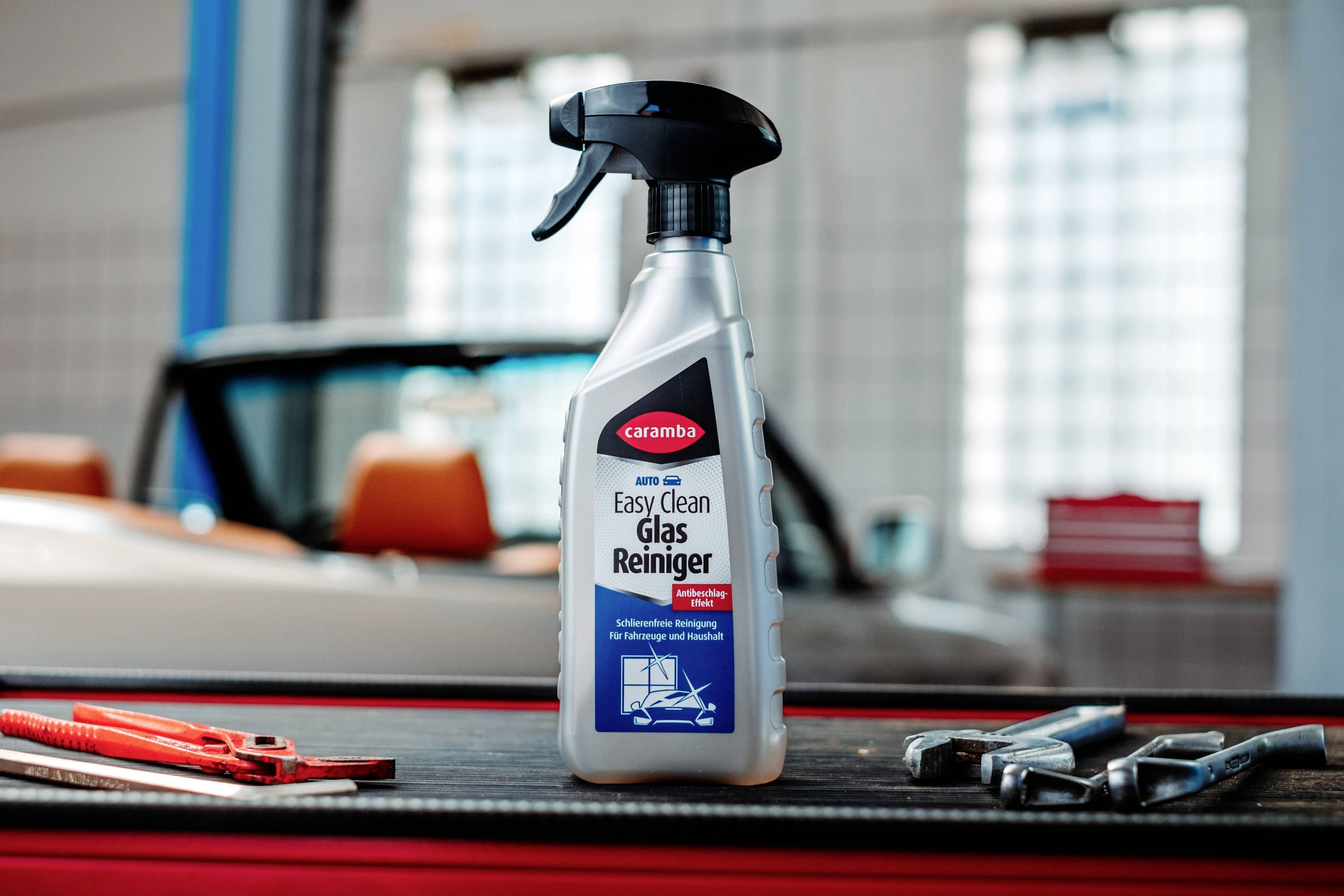 A bottle of 'Easy Clean Glas Reiniger' cleaner on a table, with car repair tools nearby and a car in the blurred background.
