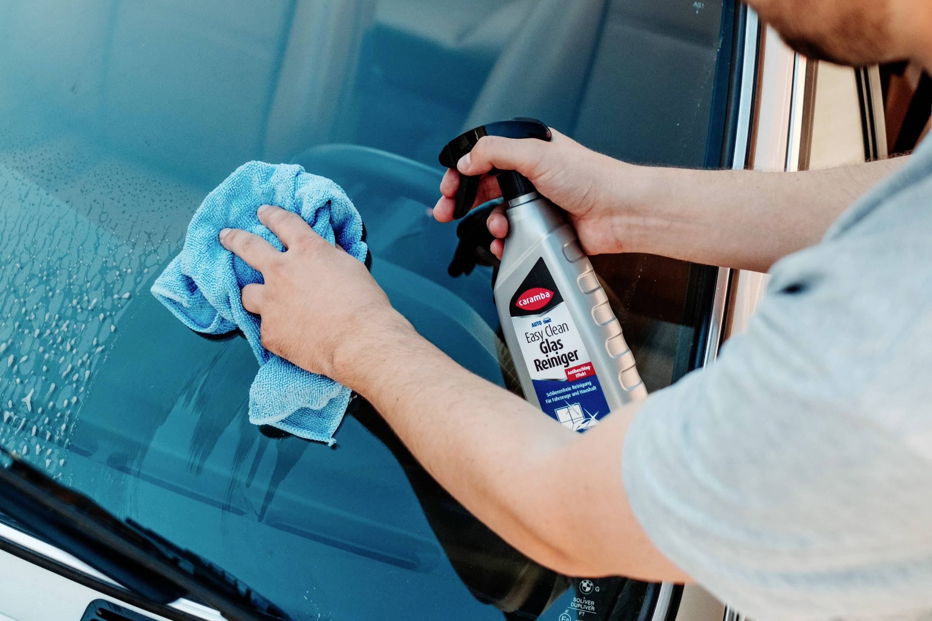 A person cleans a car windshield using a spray bottle of glass cleaner and a blue cloth.