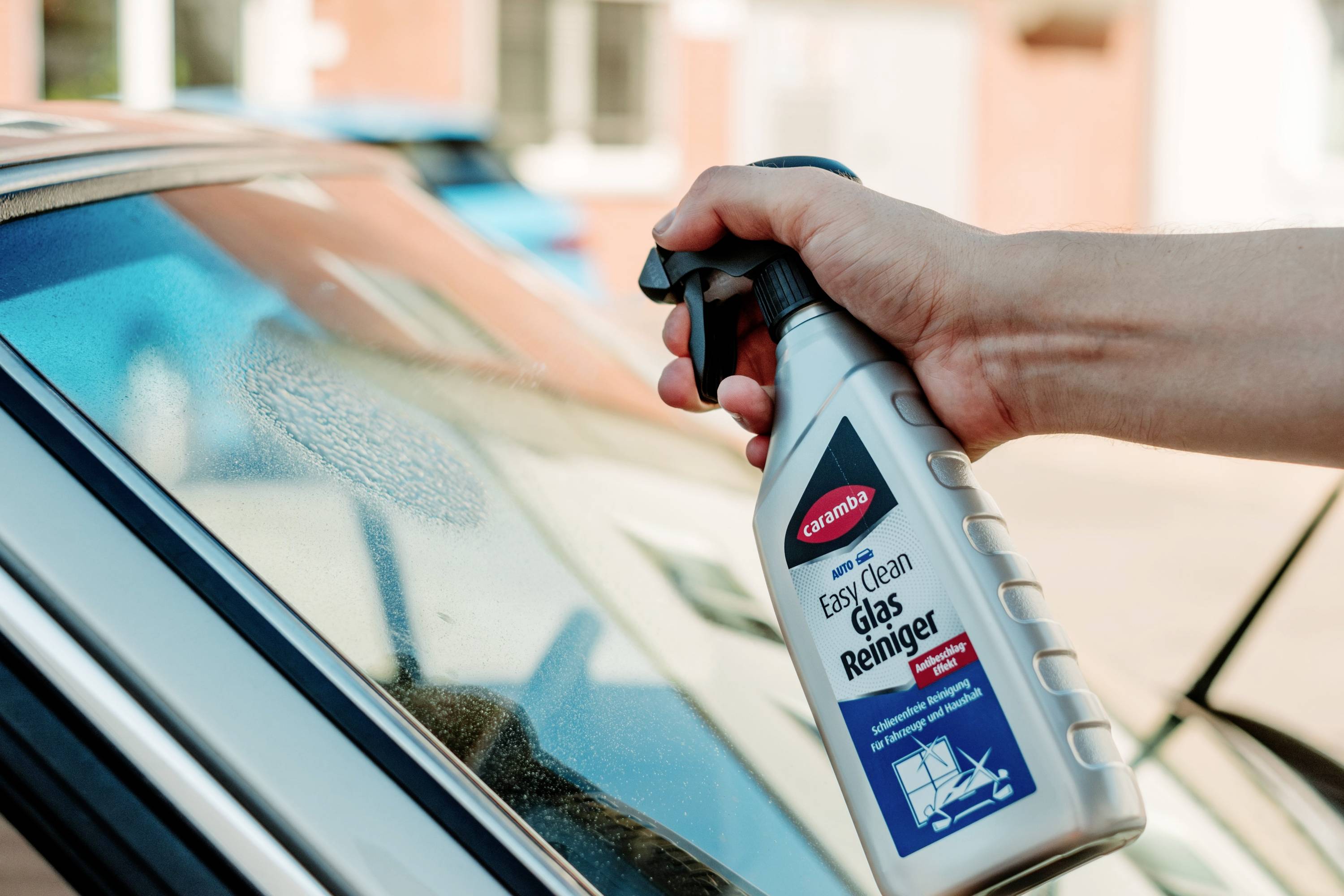 A hand sprays glass cleaner from a bottle labeled 'Easy Clean Glas Reiniger' onto a car window, creating a foamy area on the glass.