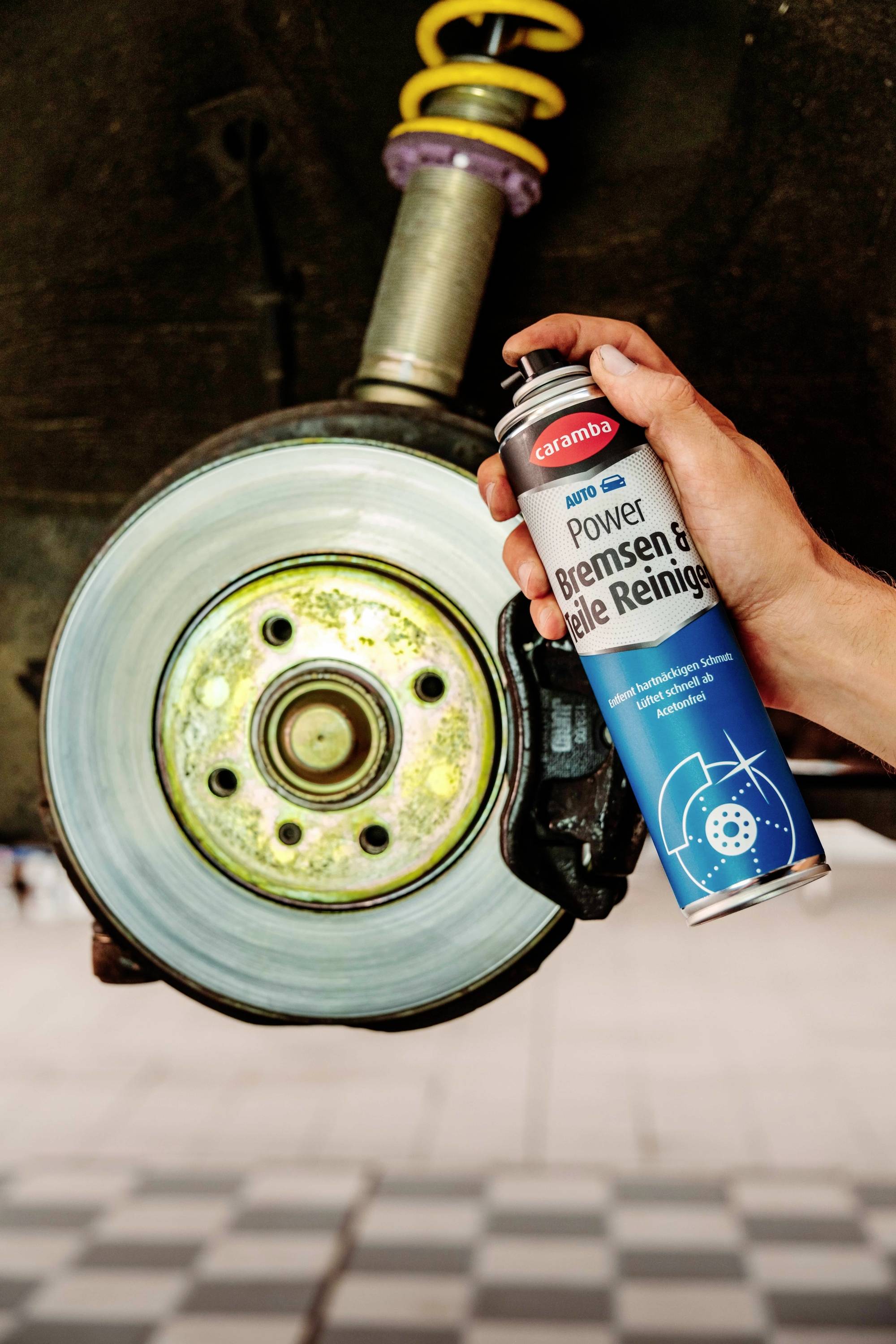 A hand sprays brake cleaner onto a car's brake disc, which is mounted on a vehicle lift, highlighting maintenance and cleaning.