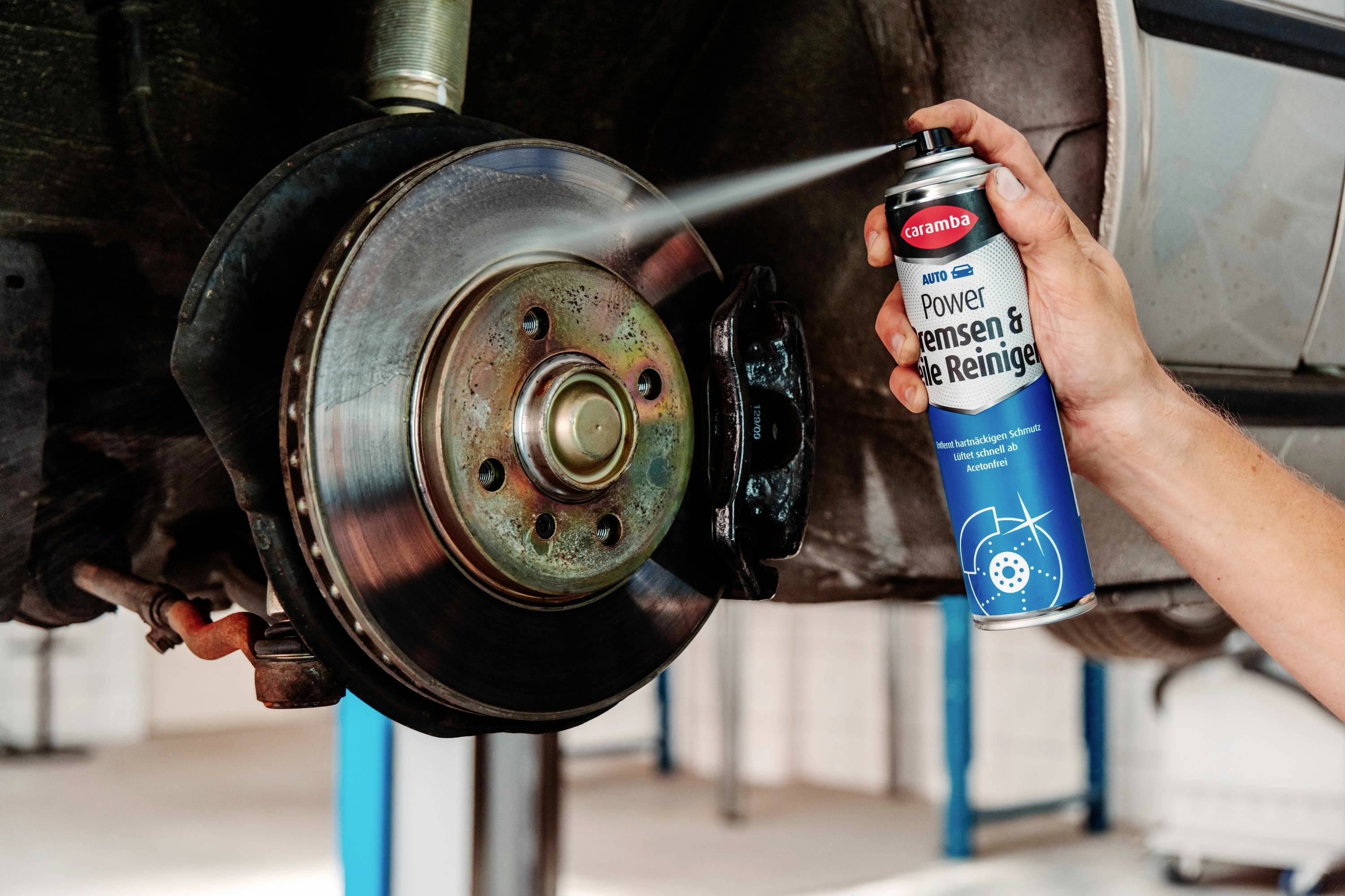 A hand sprays brake cleaner onto a car's brake disc, in a garage setting, illustrating maintenance or repair work on the vehicle.