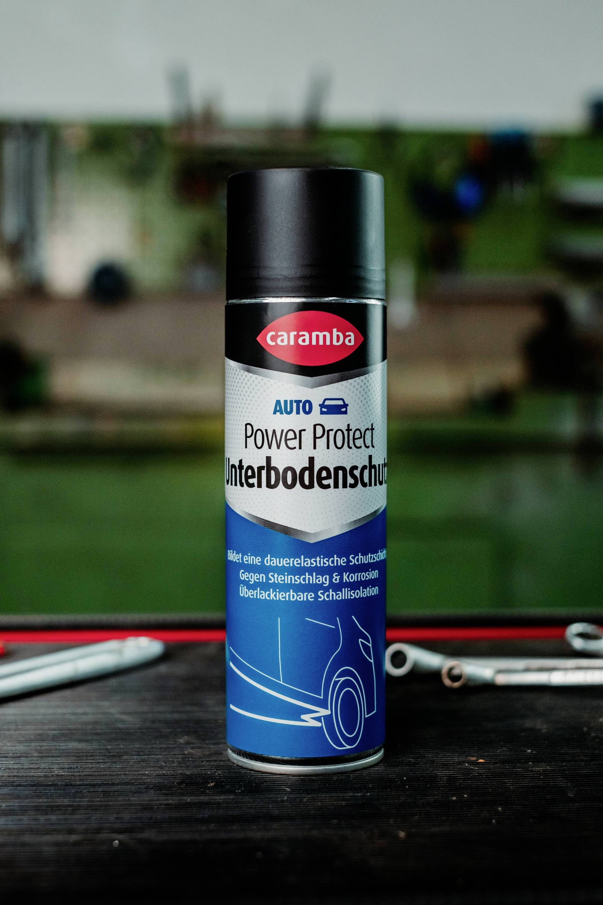 A can of Caramba Auto Power Protect underbody protection spray on a workbench with tools in a garage setting.