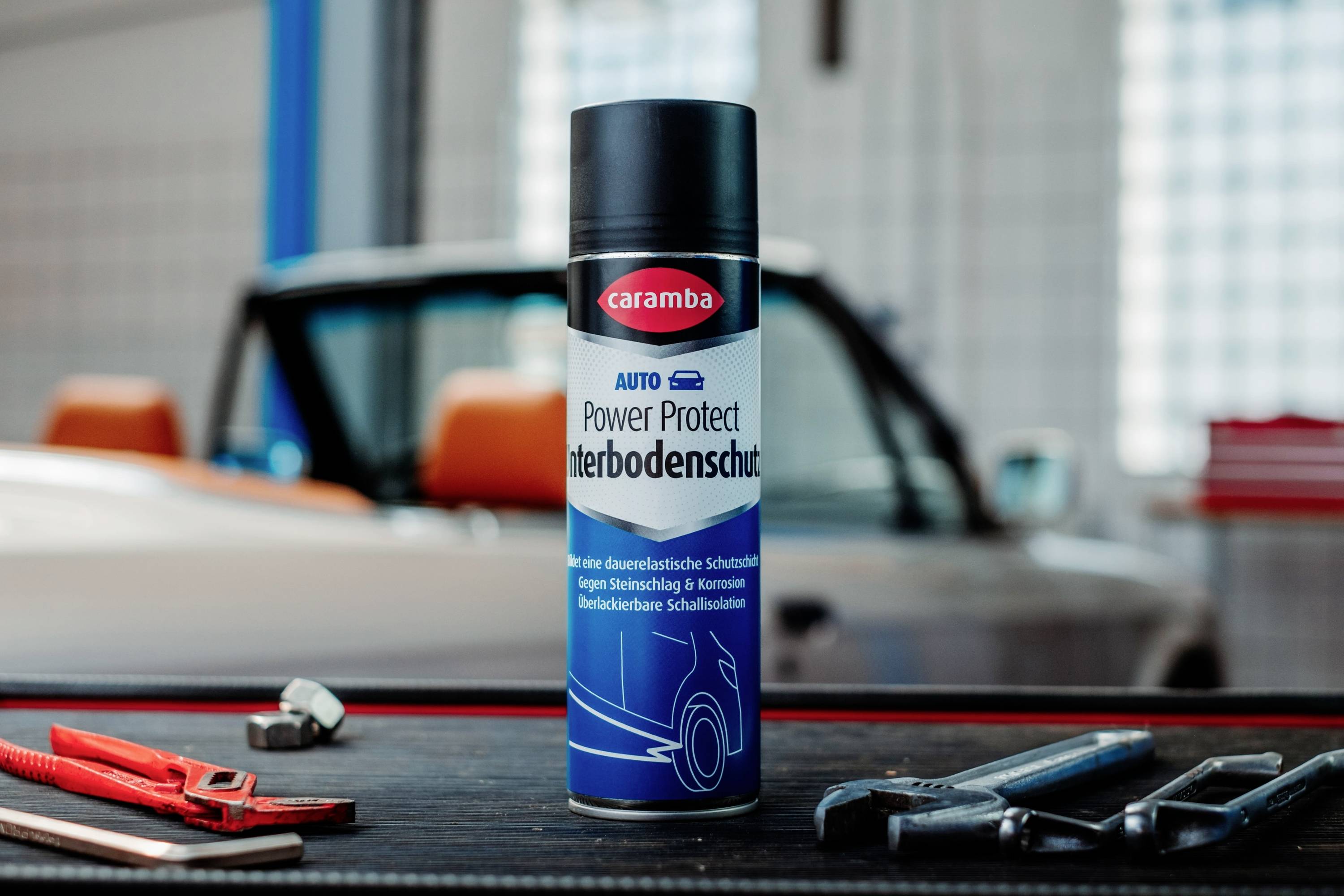 A can of 'Caramba Power Protect Unterbodenschutz' rust protection spray stands on a table with tools, in front of a classic car.