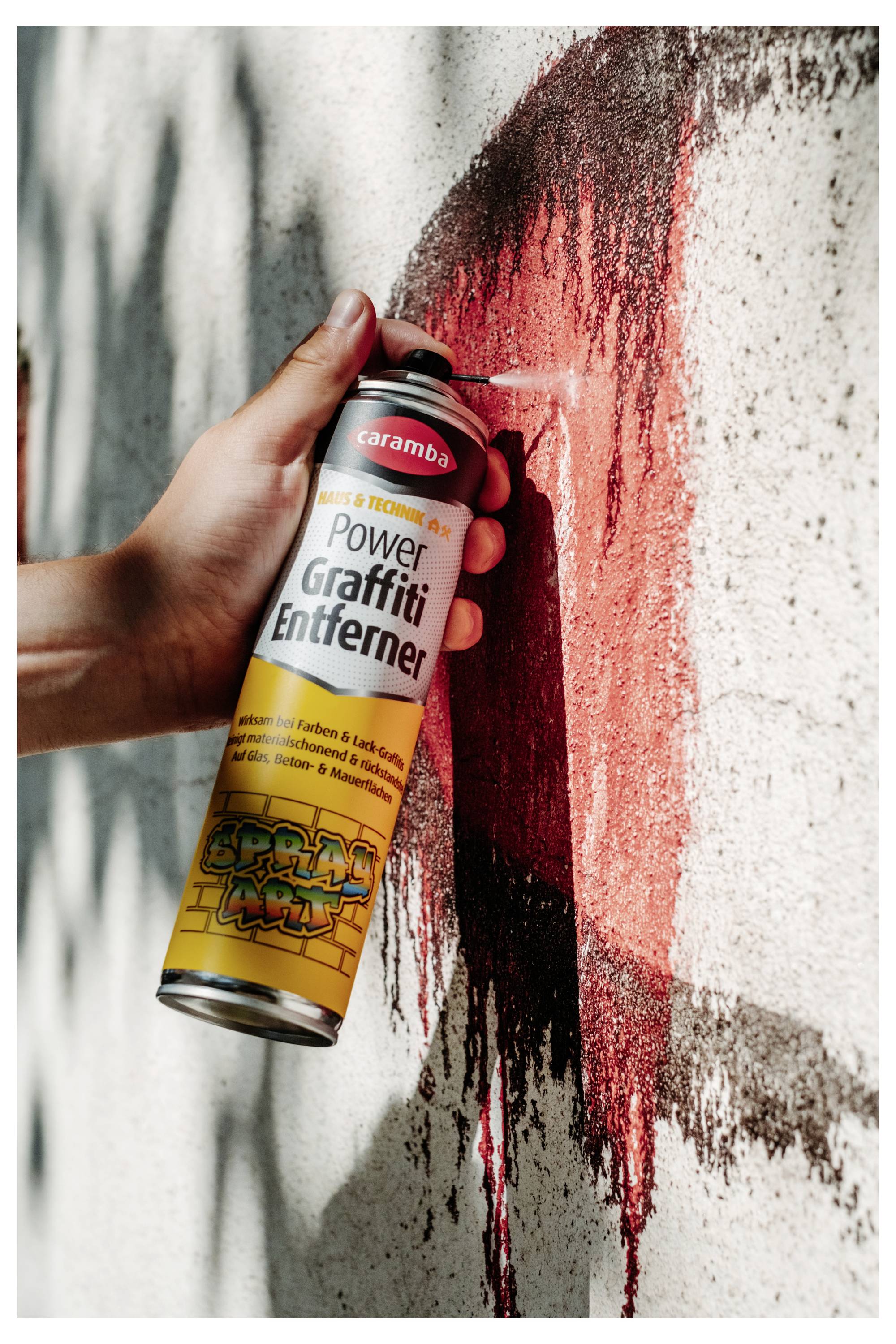 A hand sprays red paint from a 'Power Graffiti Entferner' can onto a wall, with red streaks running down the surface.