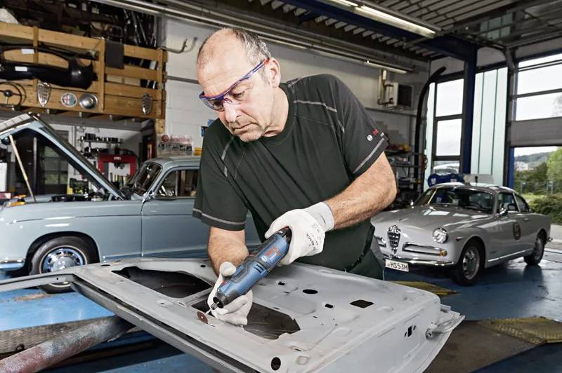 A mechanic works concentratedly in a workshop with a drill on a car part; a vintage car in the background.
