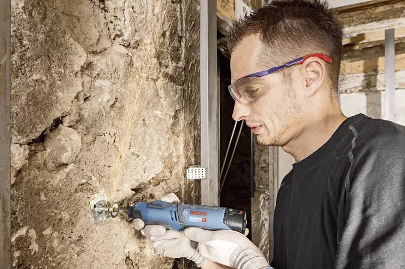A man wearing safety glasses and gloves is working with a blue Bosch multi-tool on an old stone wall.