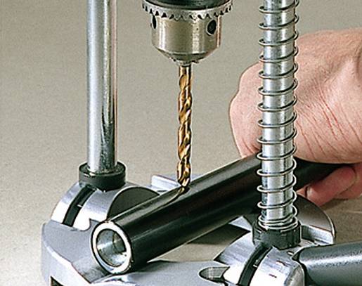 A hand is holding a metal pipe while a drill bores a hole into the pipe.