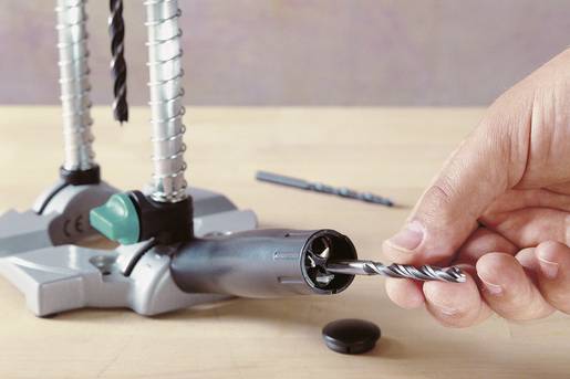 A hand is inserting a drill bit into a drill press that is positioned on a table. The focus is on the precision of the insertion.