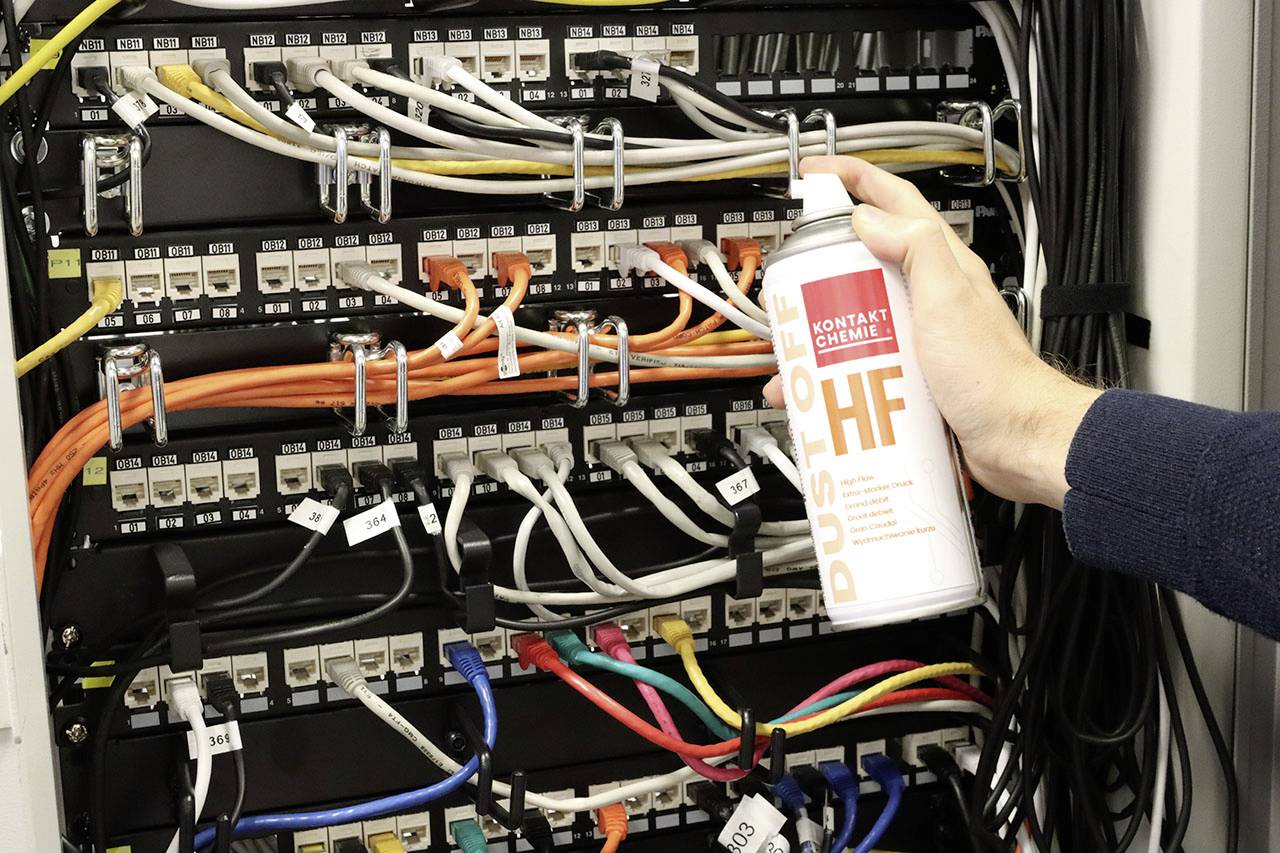 A person is holding a spray can product in front of a network server with many colourful cables to remove dust.