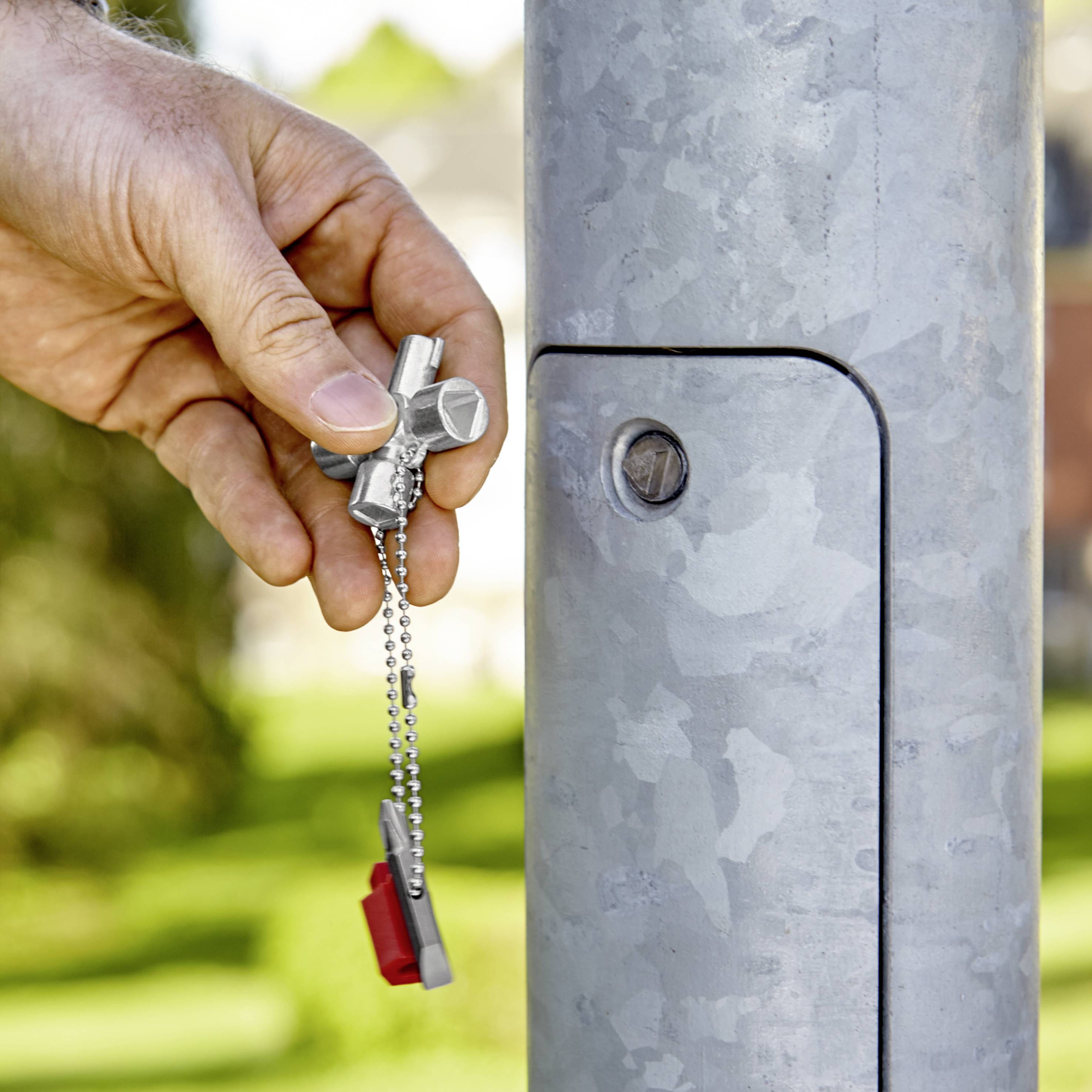 A hand is inserting a key into a lock on a silver metal post, surrounded by a green outdoor background.